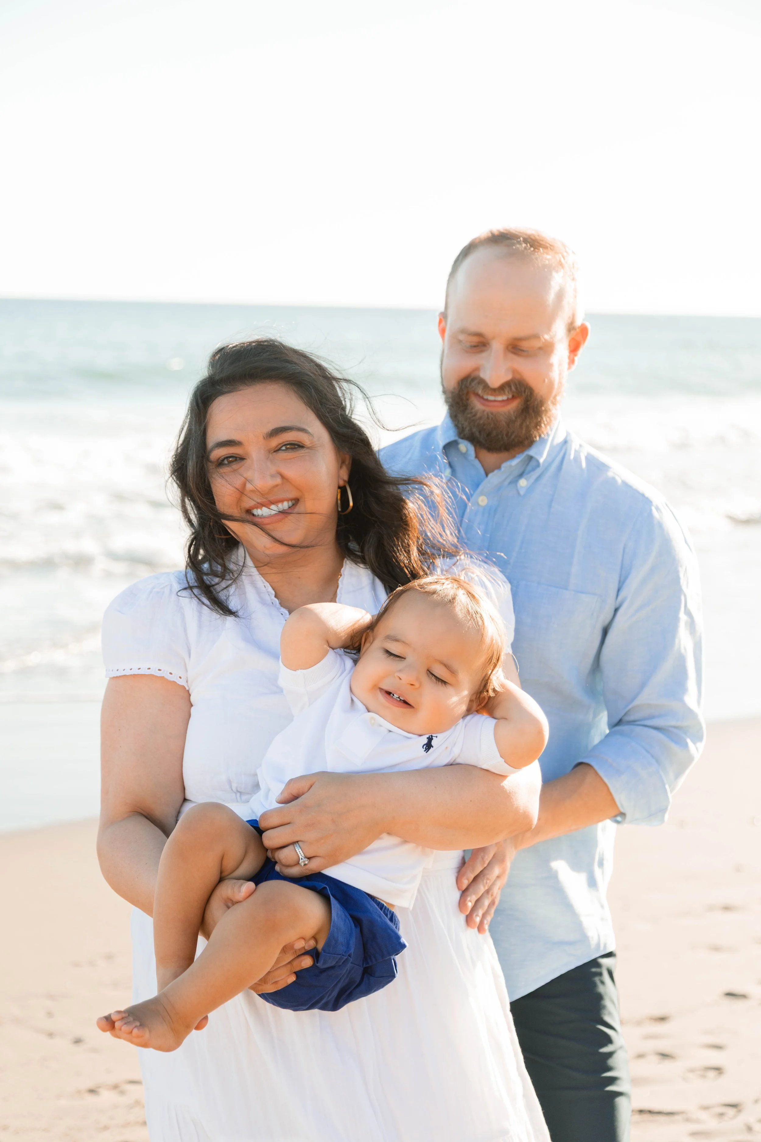 A happy family of three on the beach, with a woman holding a smiling young boy and a man standing behind them, all dressed in light-colored clothing.