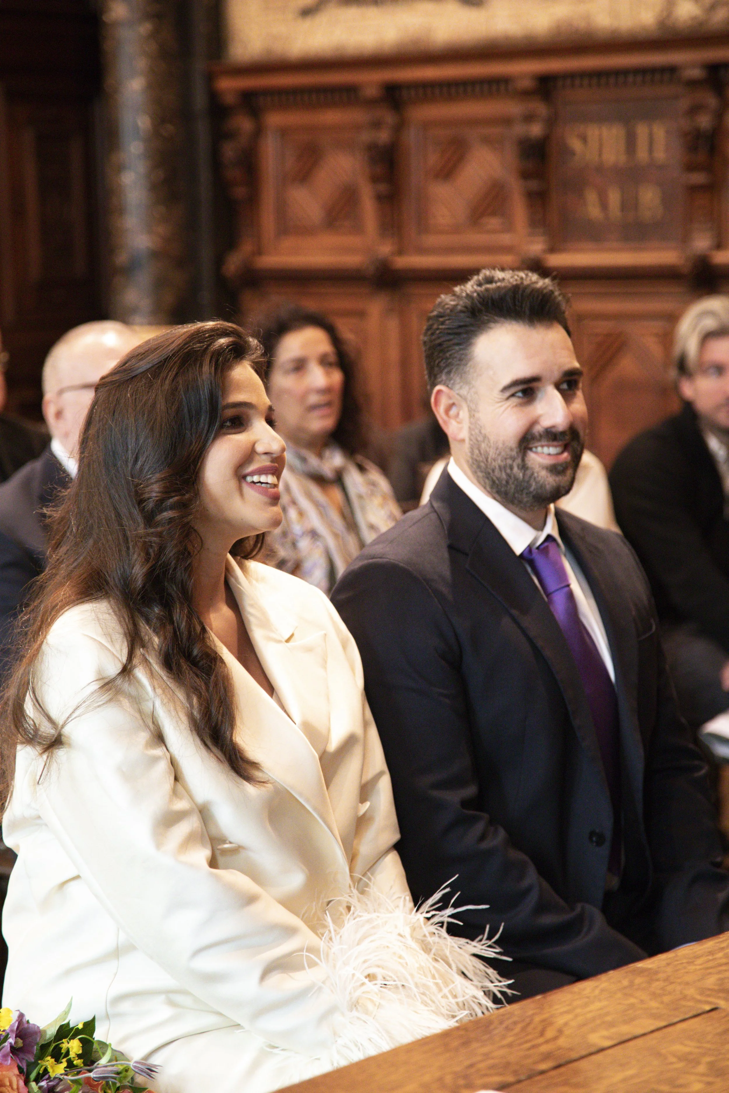 A group of people seated in an ornate wood-paneled room, smiling and dressed in formal attire. The focus is on a woman with long brown hair in a white outfit and a man with dark hair and a beard in a suit with a purple tie. Other individuals are visi