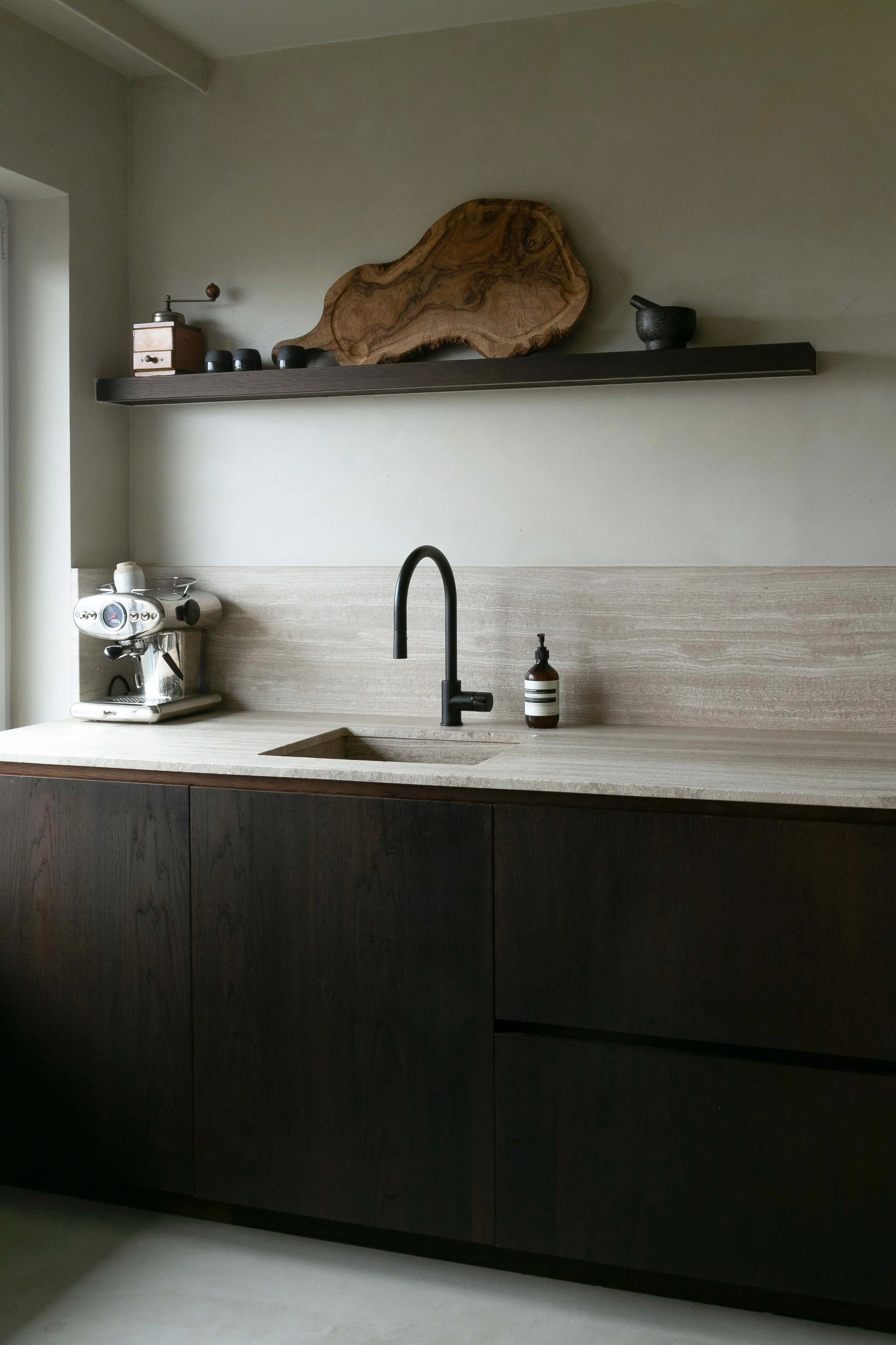Minimalist kitchen with dark lower cabinets, light countertop, and a black sink faucet; upper shelf with a wooden tray, black cups, and a black mortar and pestle; on the countertop, a silver espresso machine and a brown bottle.