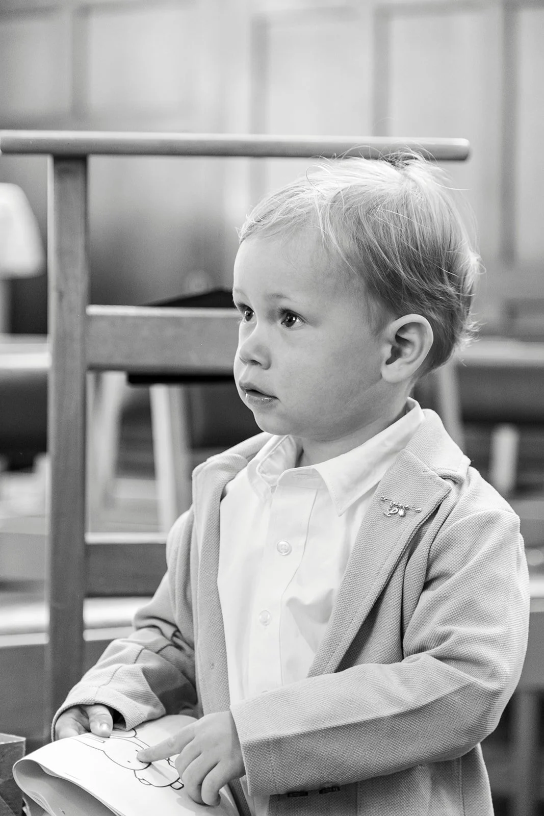 A young boy with light-colored hair, wearing a white shirt and light-colored blazer, sitting indoors, holding a coloring book and pointing at a picture.
