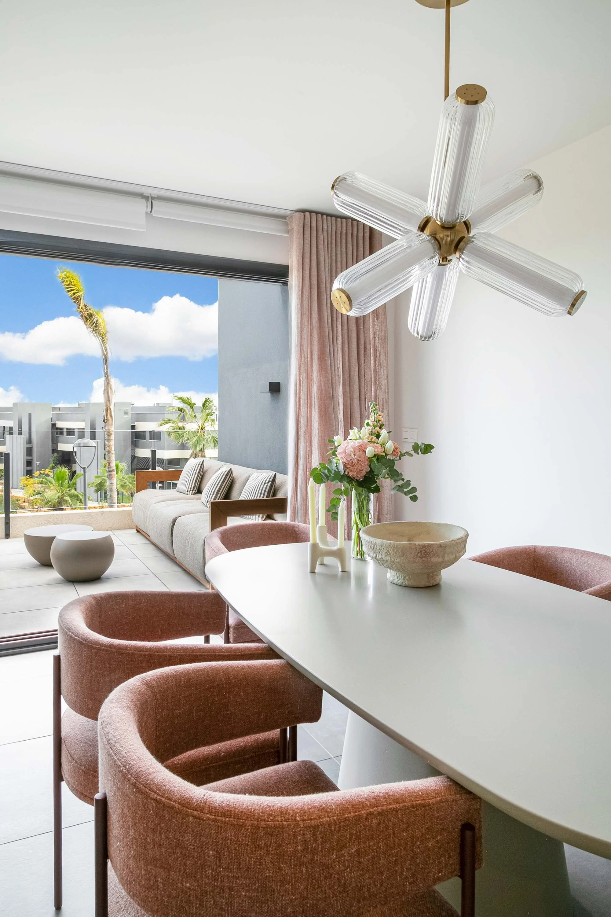 A modern living and dining room interior with pink chairs around a white table, a floral arrangement, and a view of a balcony with palm trees outside.