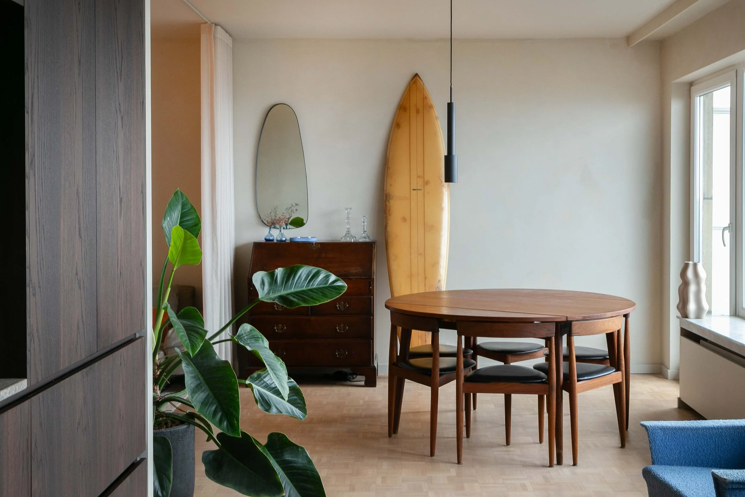A cozy, sunlit dining area with a wooden table and four matching chairs, decorated with a surfboard and a mirror on the wall, and a large green plant in the foreground.