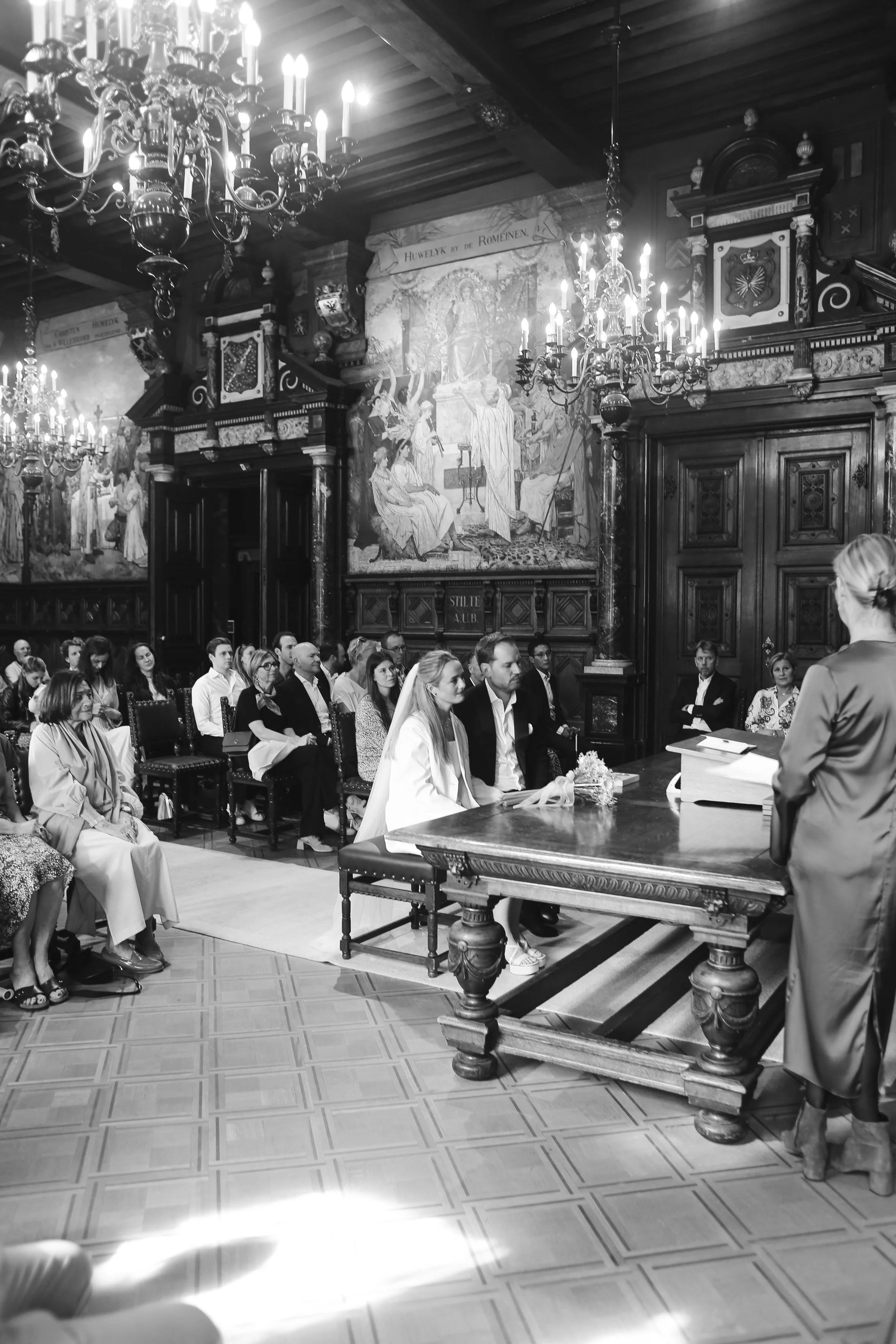 A formal gathering in a large, ornate room with high wooden ceilings, grand chandeliers, dark wood paneling, and large wall murals. People are seated at long tables, listening to a woman standing and speaking at a table with documents and flowers.