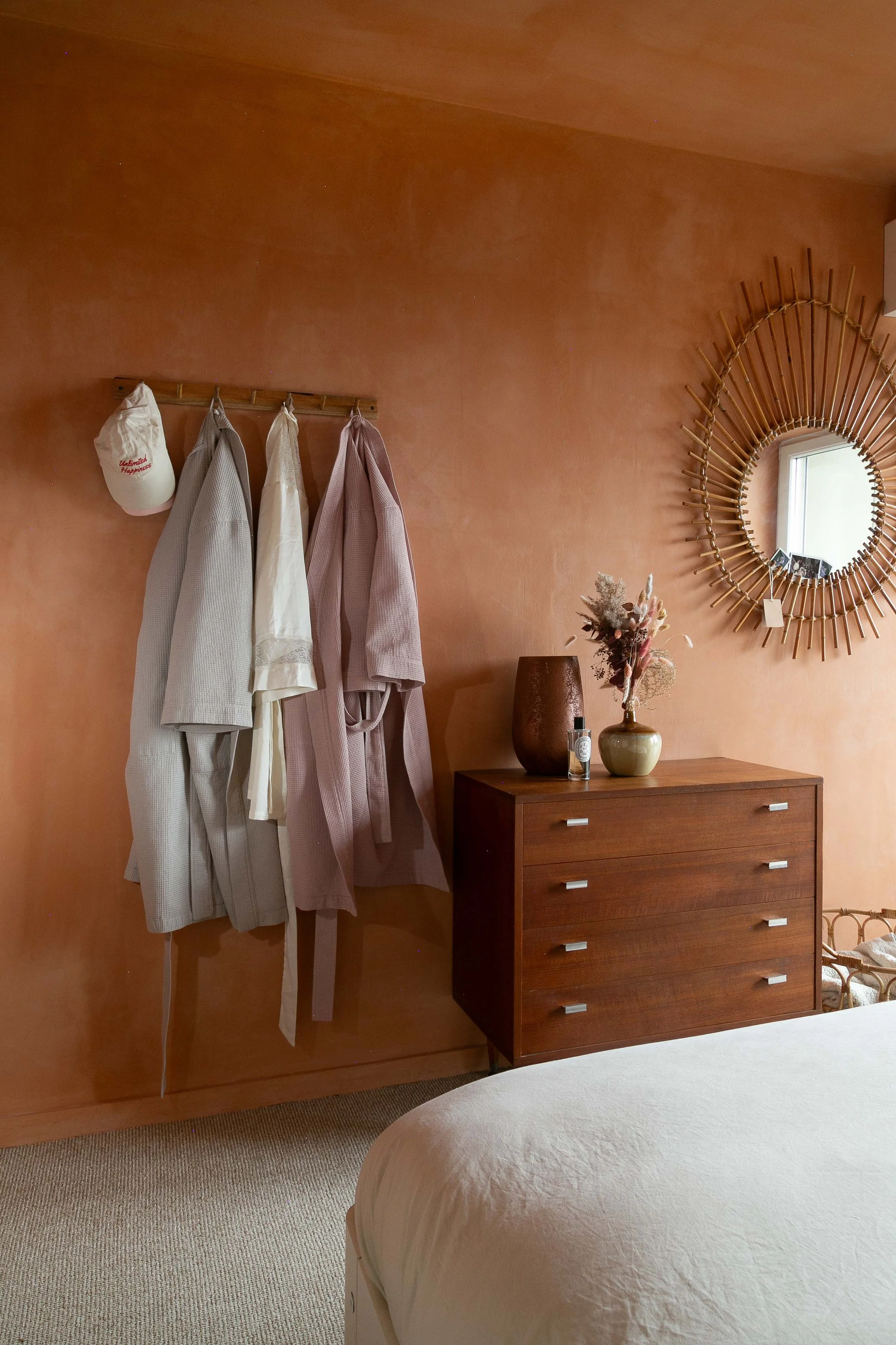 A bedroom with a peach-colored wall. On the wall, a wooden coat rack holds three bathrobes and a white baseball cap. To the right, a round mirror with a sunburst frame hangs above a wooden dresser with four drawers. The dresser has vases and dried fl