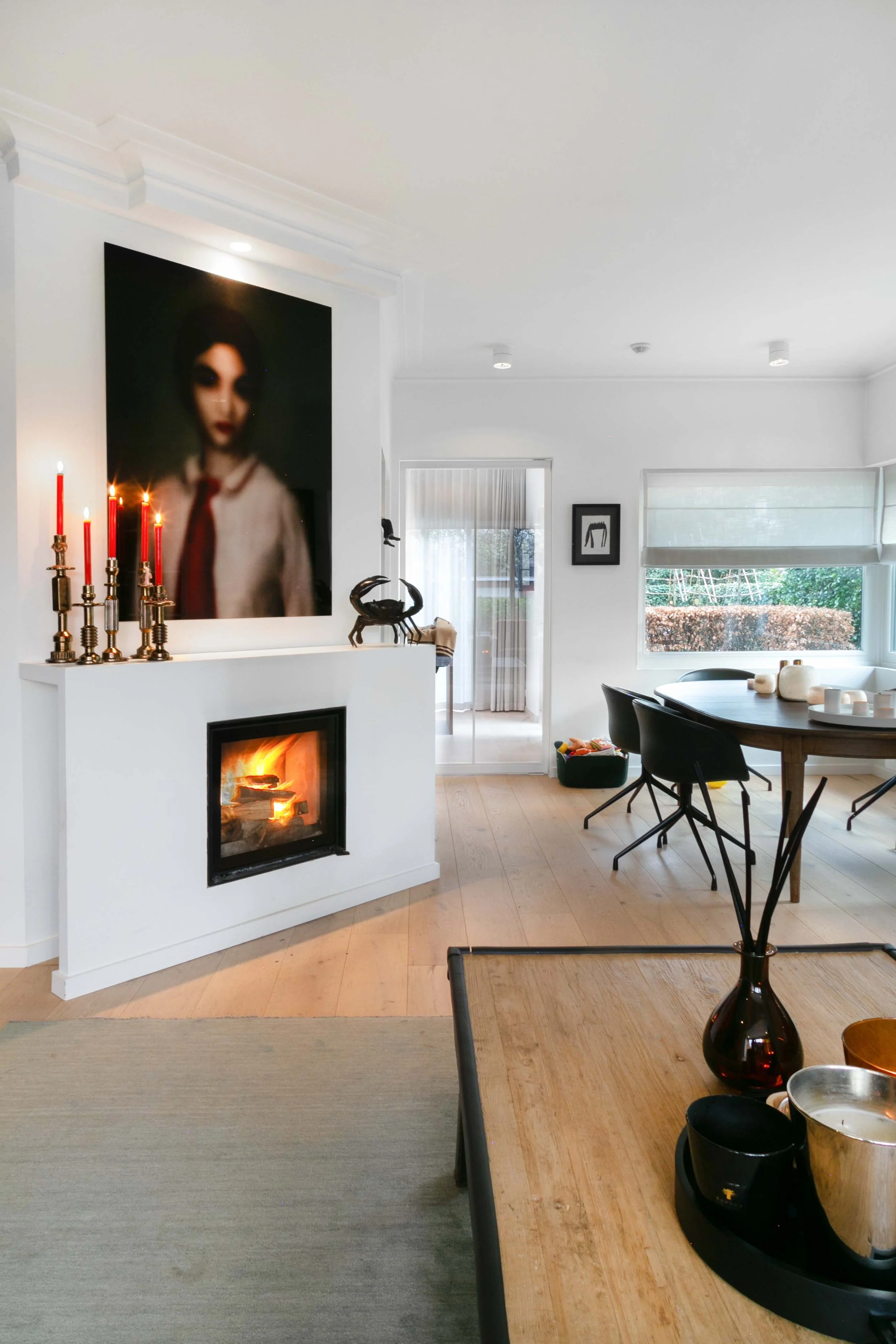 A modern living room with a white fireplace, a large abstract painting of a woman on the wall, and a dining table with black chairs. The room has large windows with white blinds, and decorative objects including a black sculpture, candles, and vases.