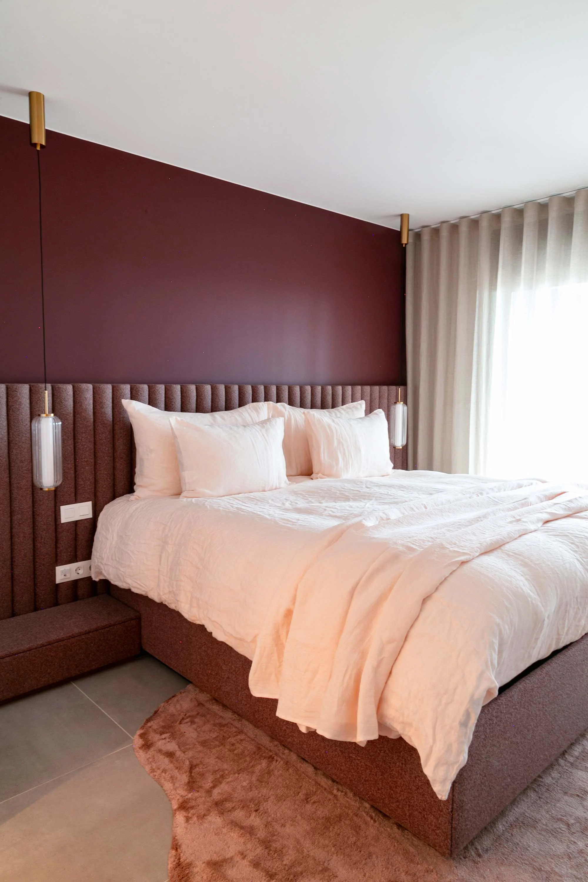 A neatly made bed with white pillows and a pale pink blanket, set against a burgundy accent wall with beige curtains and two pendant lights on either side.