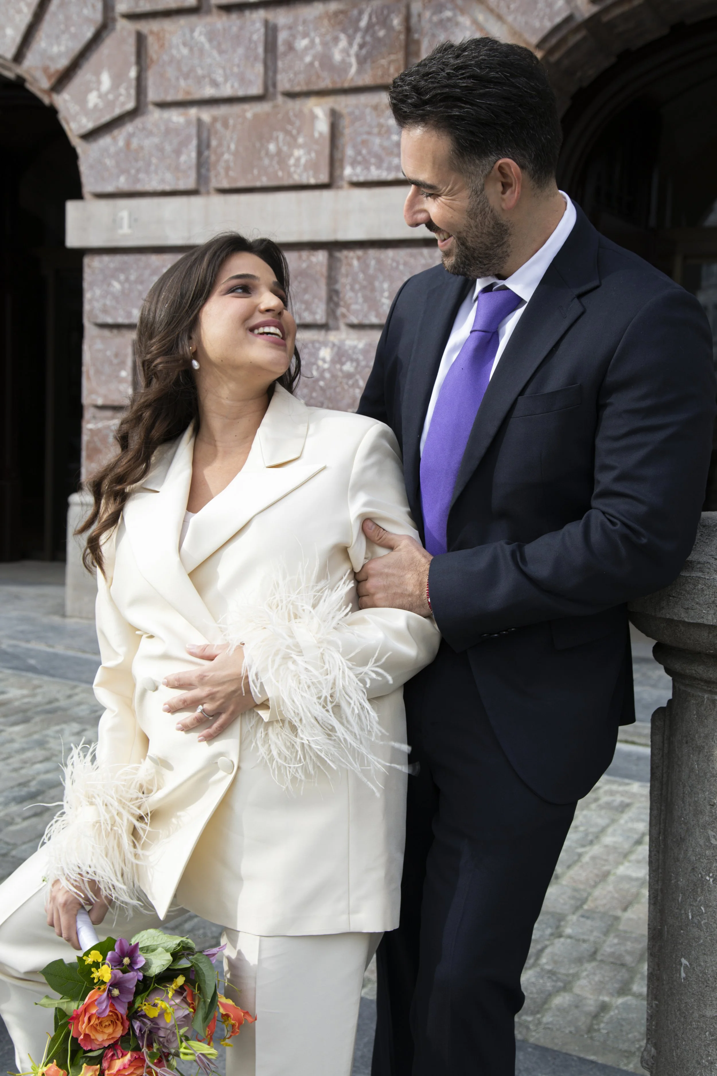 A man and woman dressed in formal attire smiling and looking at each other outside a brick building, with the woman holding a bouquet of colorful flowers.