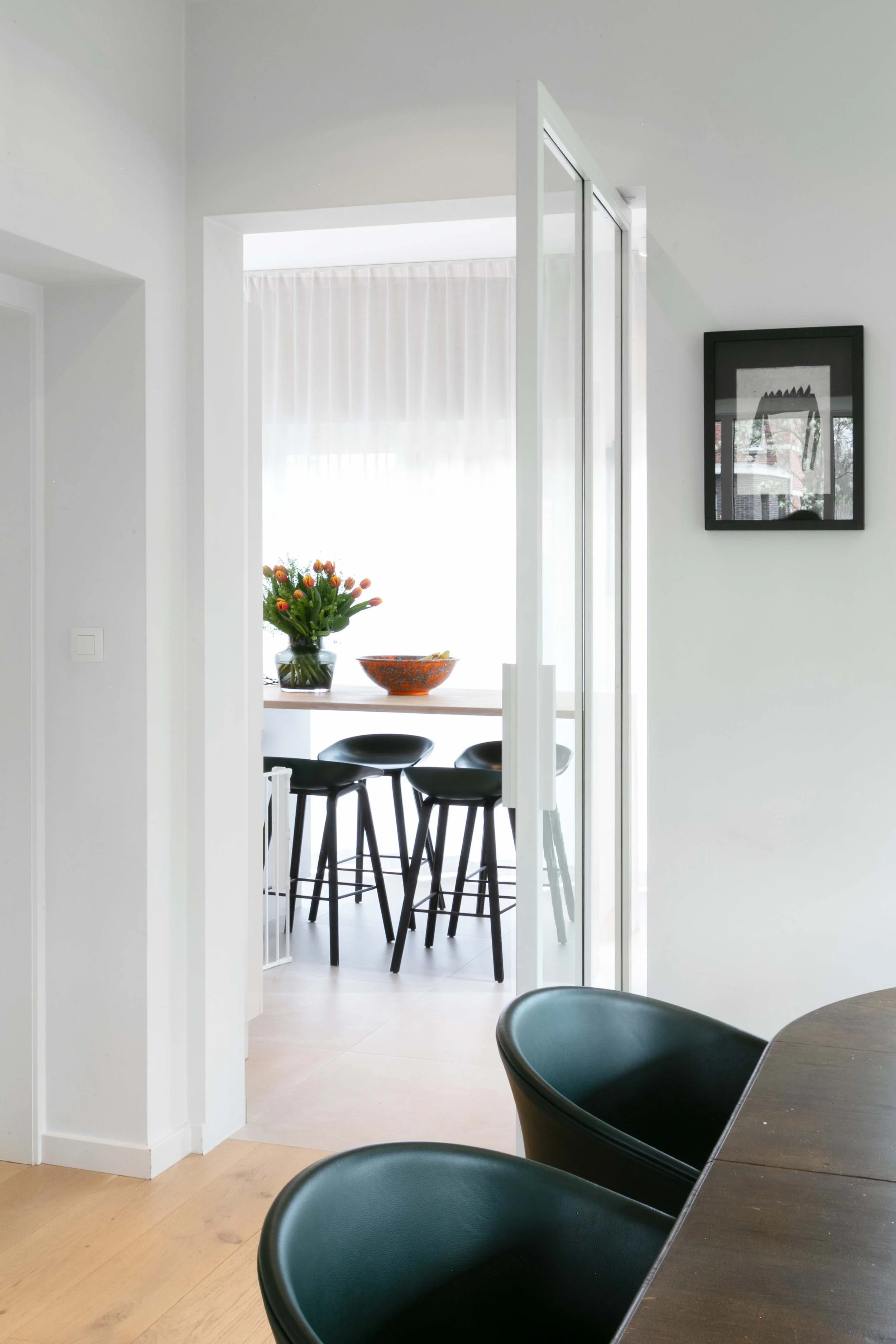 A modern dining area with black chairs around a wooden table and a kitchen in the background. There is a vase filled with tulips on the table and a framed artwork on the white wall.