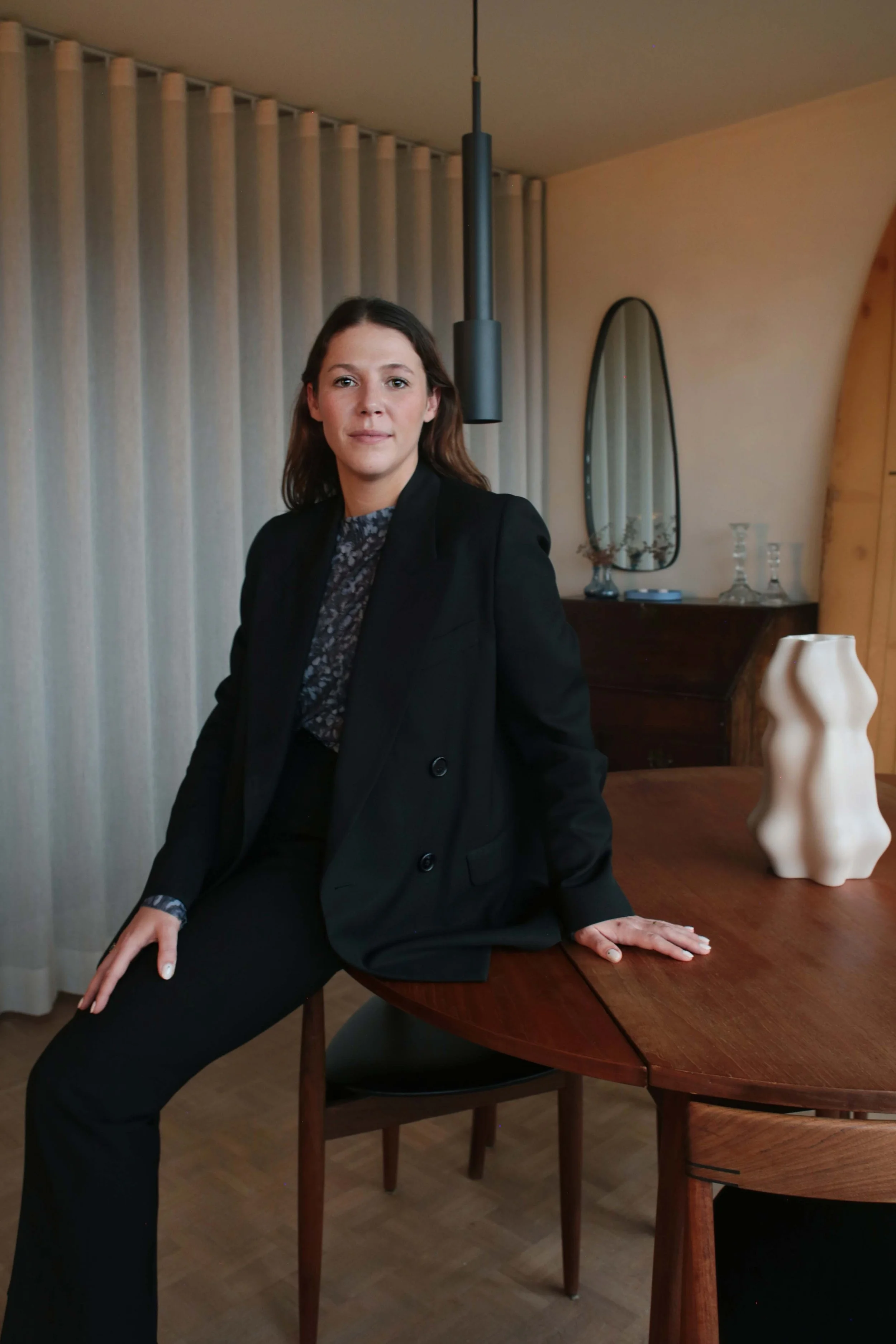 A woman with shoulder-length brown hair, dressed in a black blazer and black pants, sitting on a wooden chair with one hand resting on a round wooden dining table.