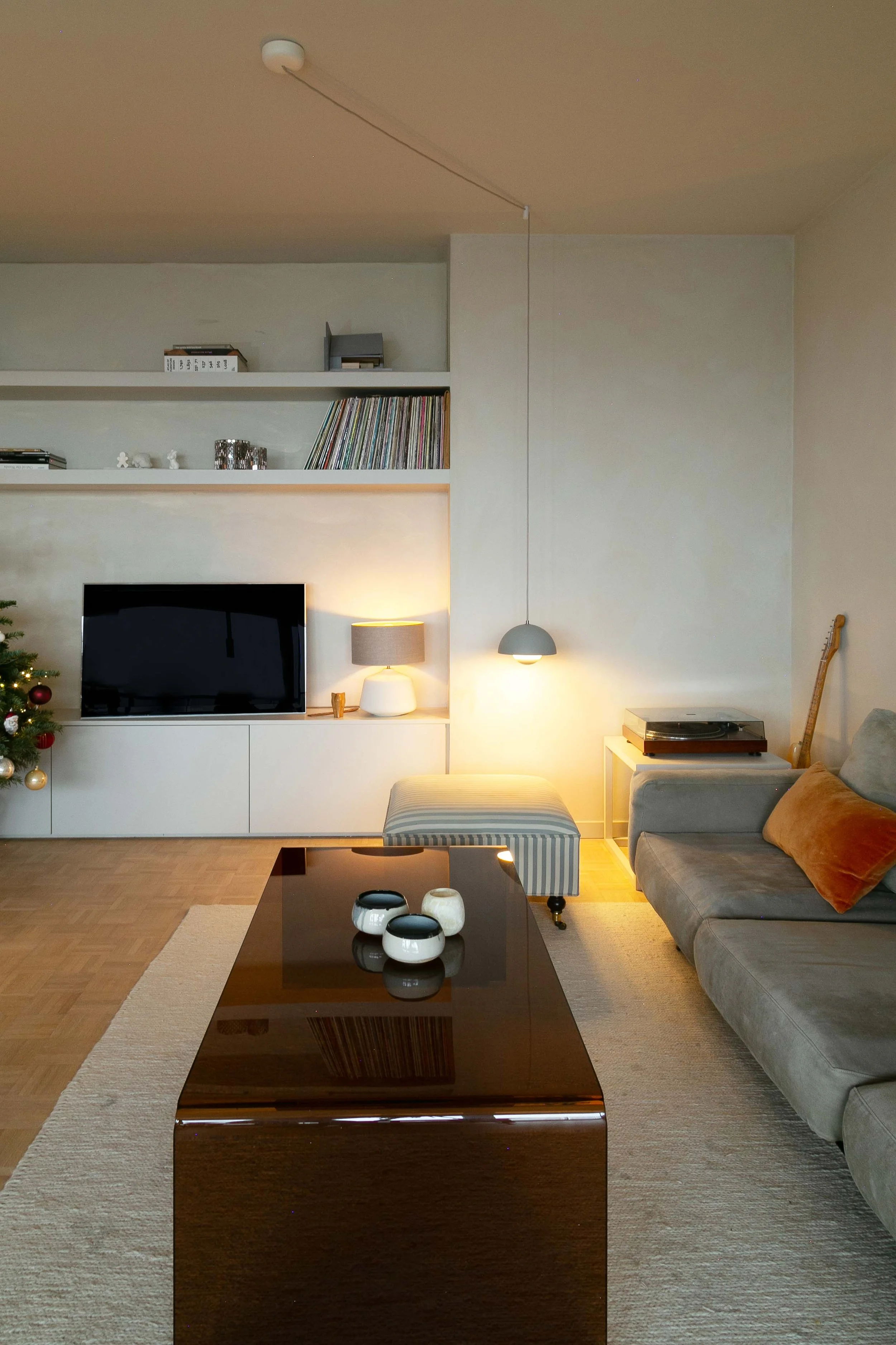 Living room with a gray sofa, a black coffee table with ceramic bowls, a TV, a beige rug, a striped ottoman, and a lamp, with a wall-mounted shelf holding records and decorative items; a wooden guitar is in the corner.