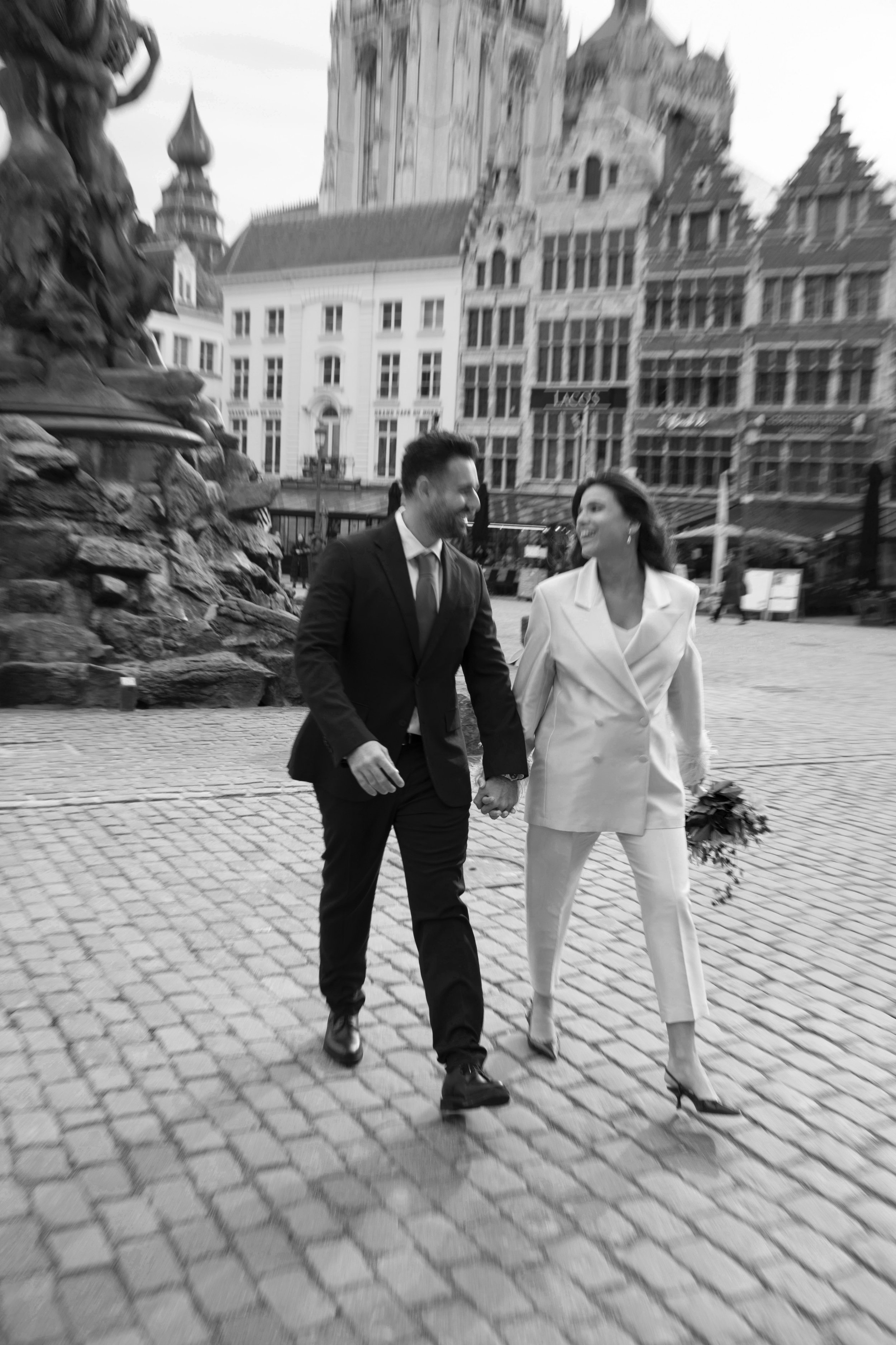 A black and white photo of a couple walking hand in hand in a European city square, smiling and looking at each other, with historic buildings in the background.