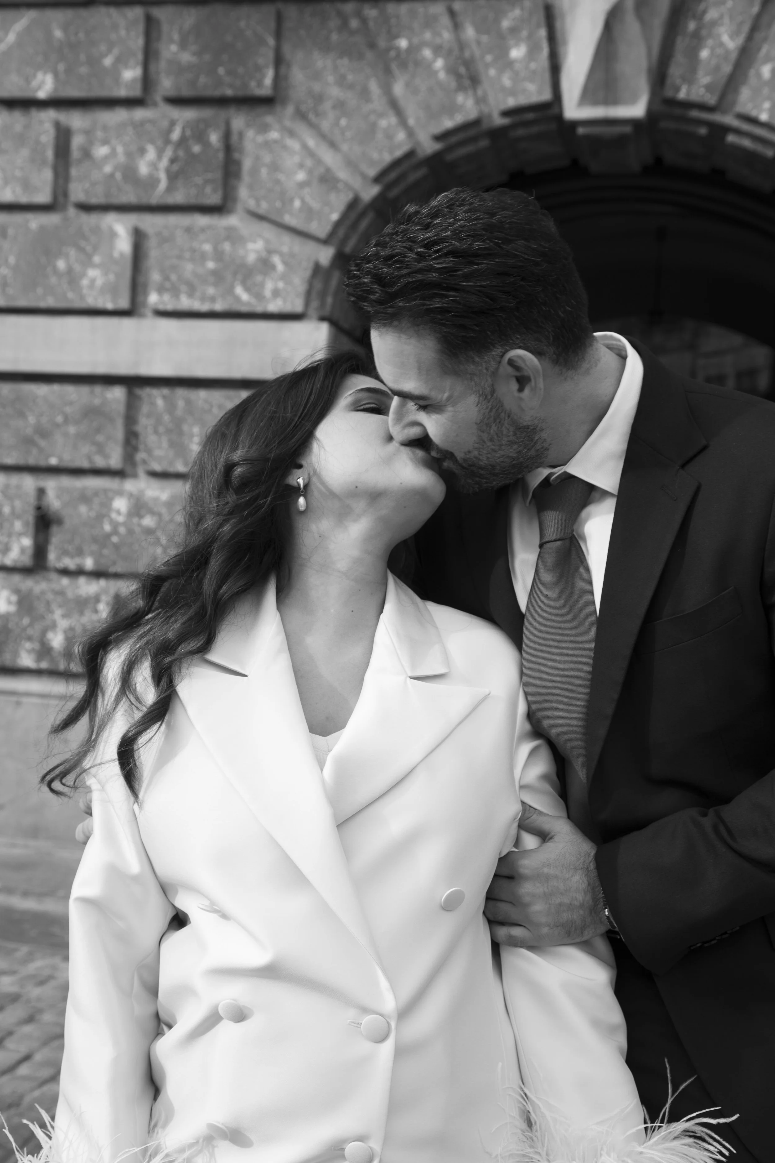 A black and white photo of a man and woman kissing closely, dressed in formal attire, in front of a brick archway.