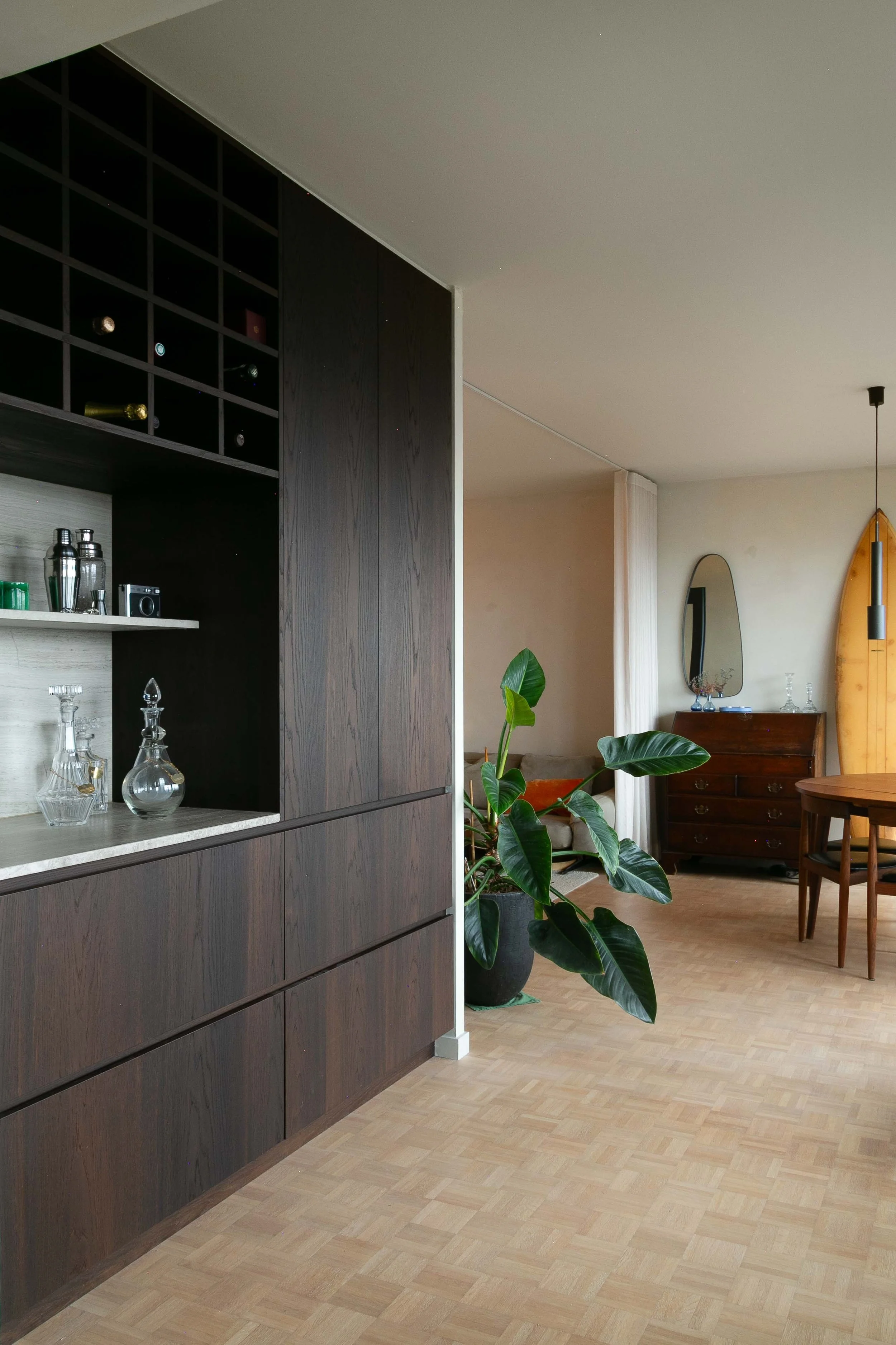 Interior view of a modern living space with a dark wooden cabinet, glass decanters, a large houseplant, a wooden dresser, and a round dining table with chairs. There is a mirror on the wall and a yellow surfboard leaning against the wall.