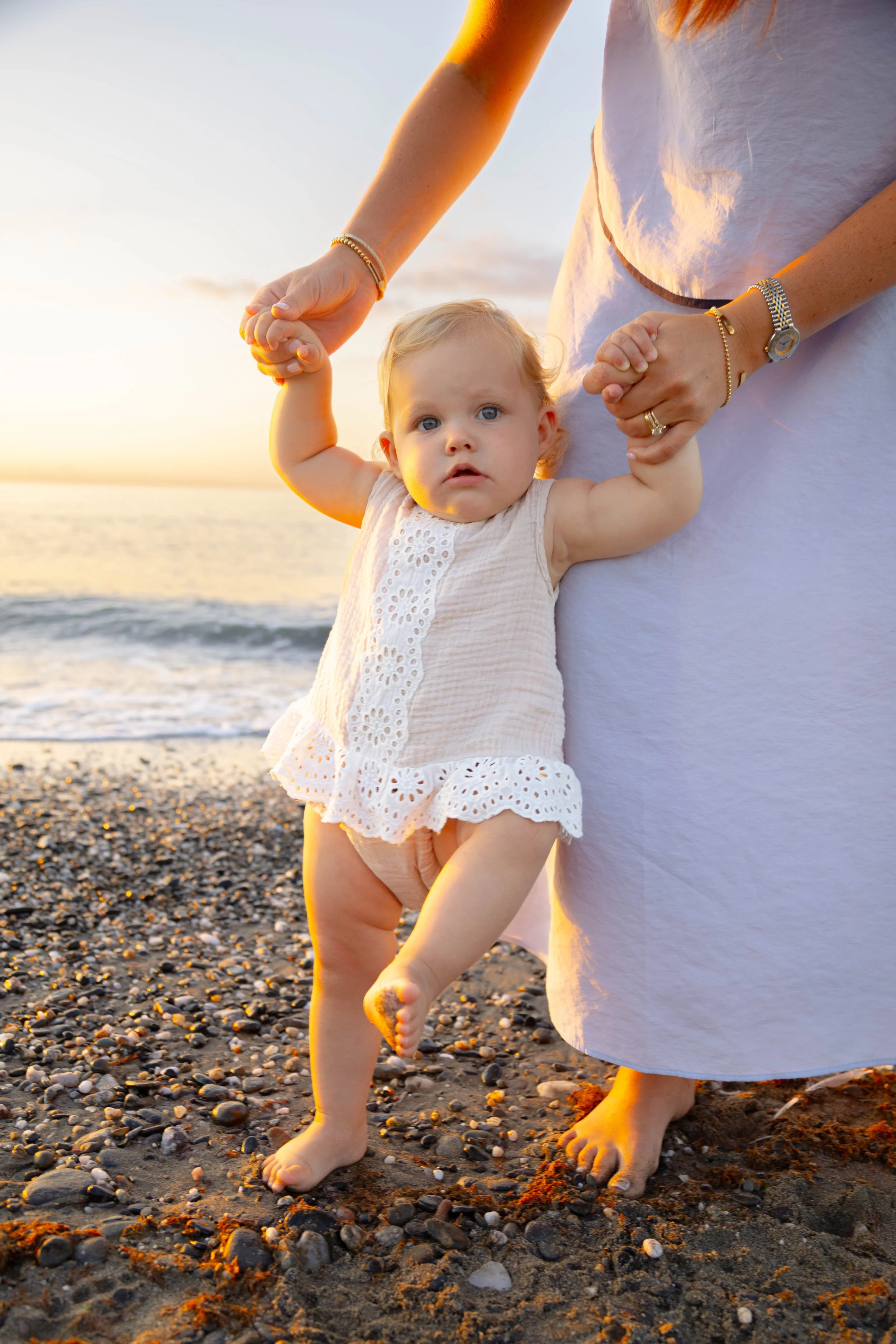 A young child in a white dress being held by an adult on a pebble beach at sunset, with the ocean in the background.