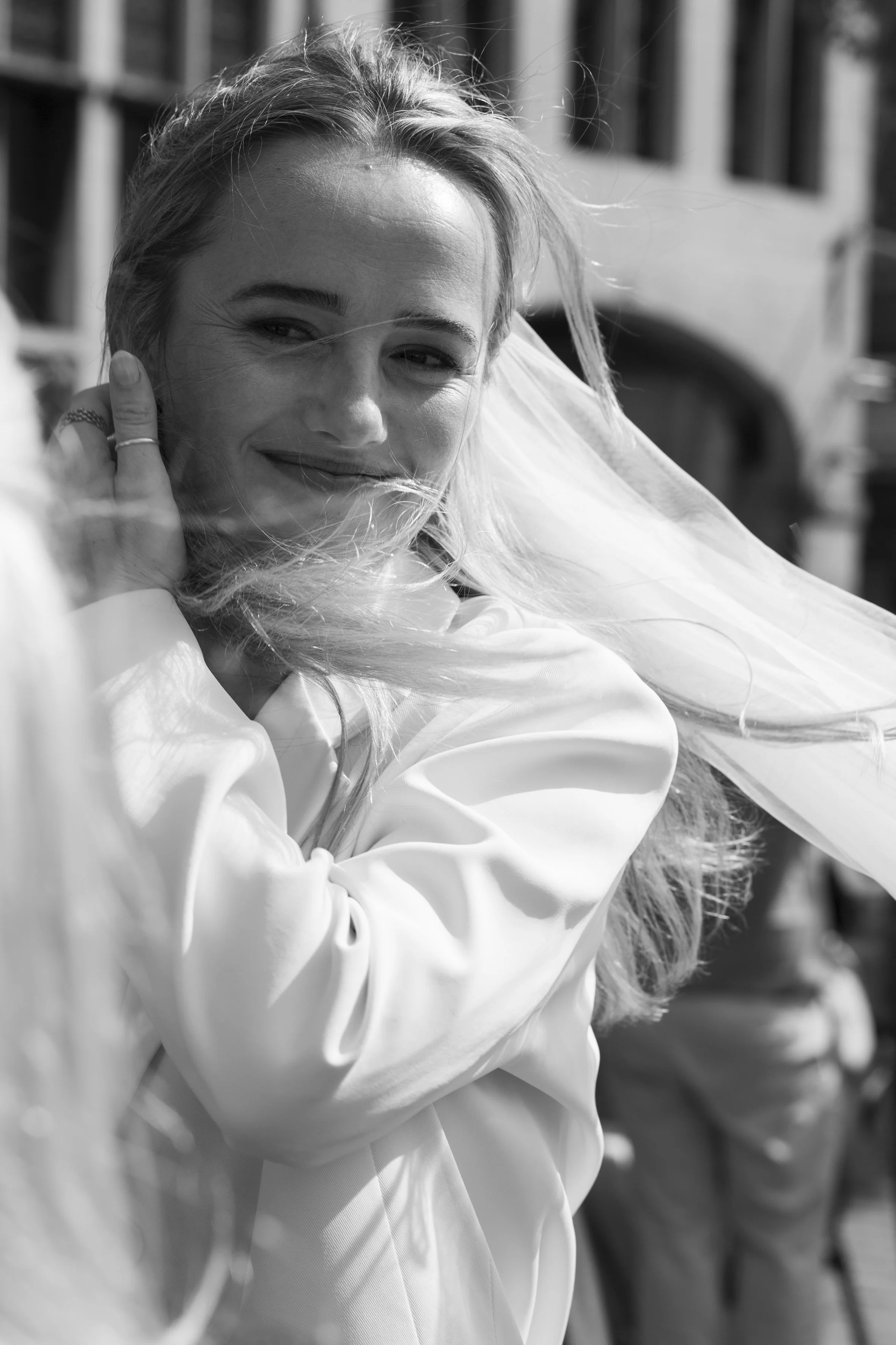 Close-up black and white photo of a smiling woman with long, flowing hair, wearing a white jacket, outdoors with a blurred background.