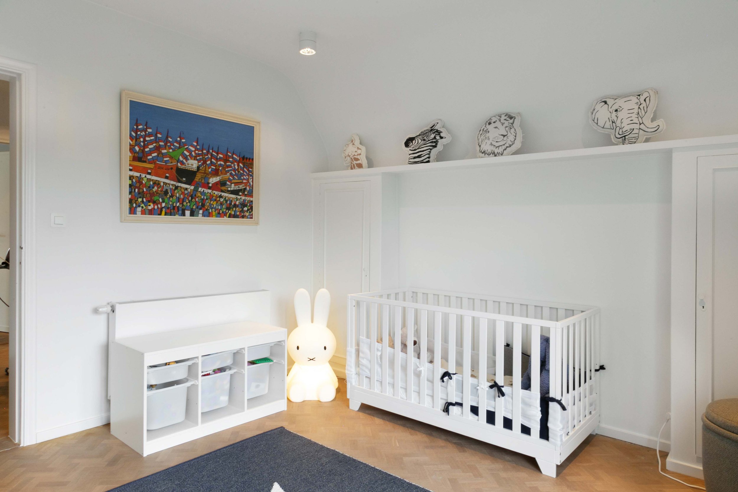 A white crib with black ribbons on the sides is placed in a nursery with light-colored wooden flooring. To the left, there is a white storage cube with small drawers, and a bunny-shaped night light is on the floor near the crib. A colorful painting d