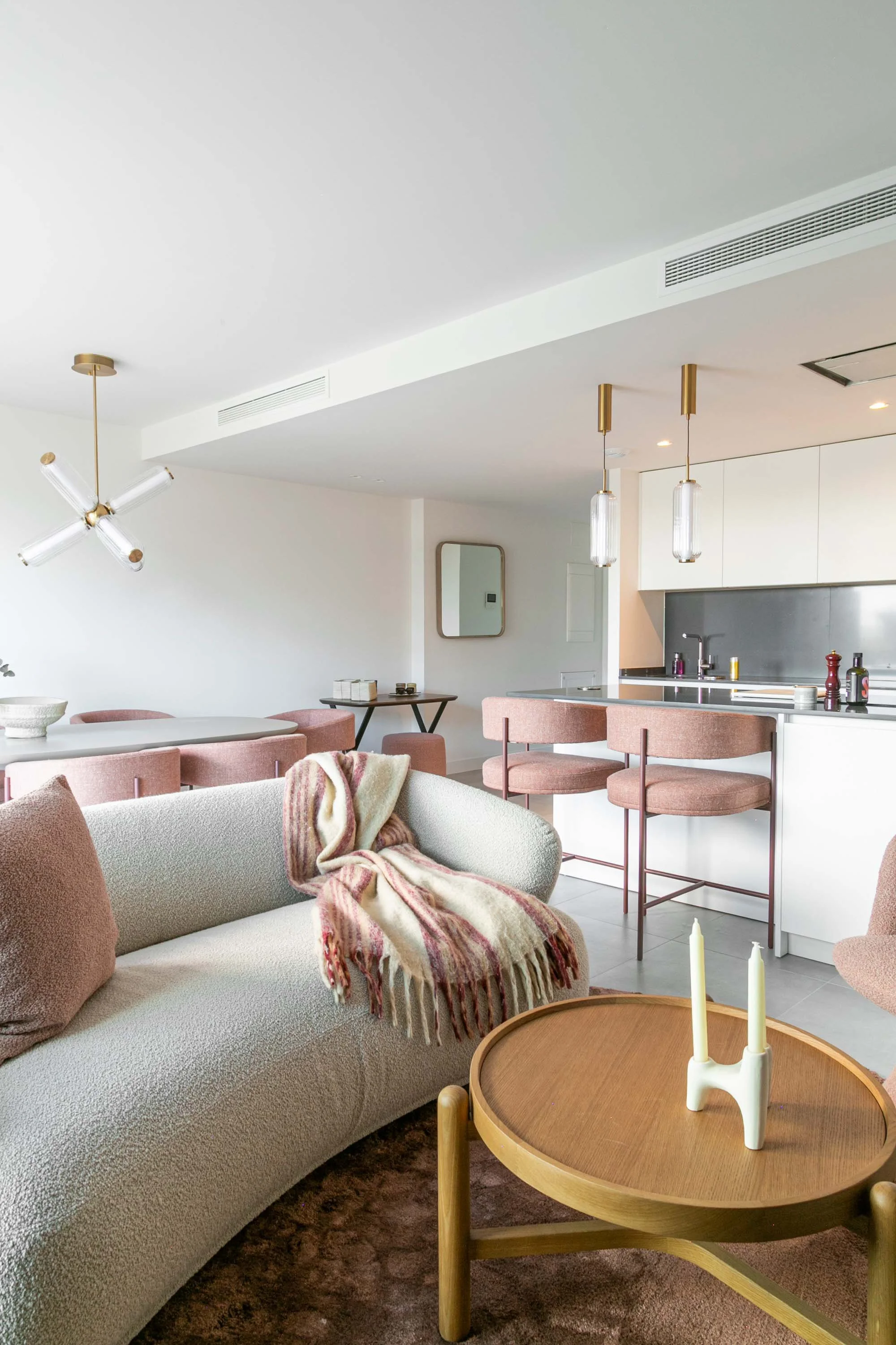 Modern living room with a beige sofa, pink chairs, a wooden coffee table with white candles, white walls, a kitchen area with hanging pendant lights, and decorative accents.