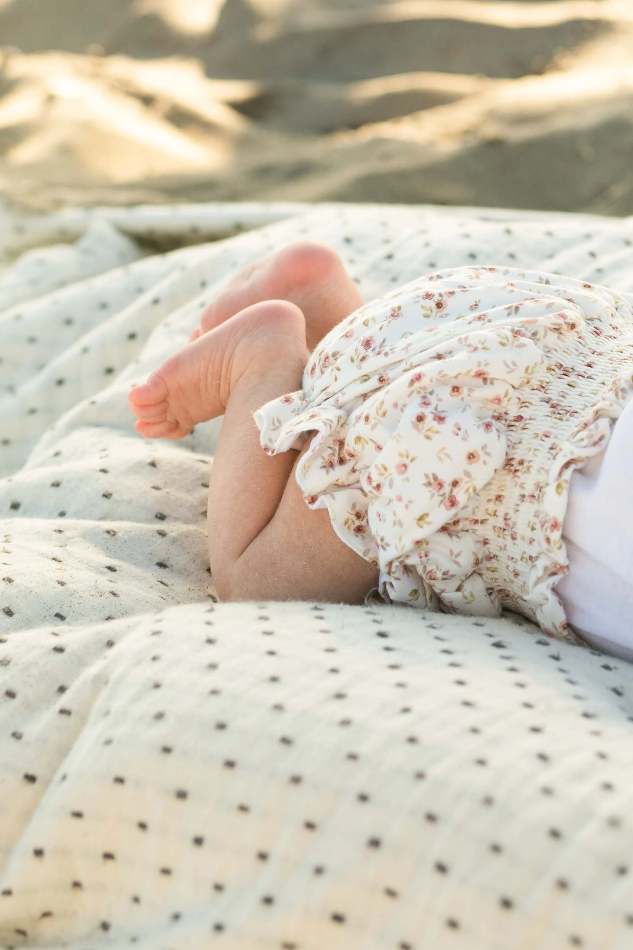 Close-up of a baby's hand resting on an adult's leg, with a white, floral, ruffled dress and a light-colored, polka-dotted blanket or clothing.
