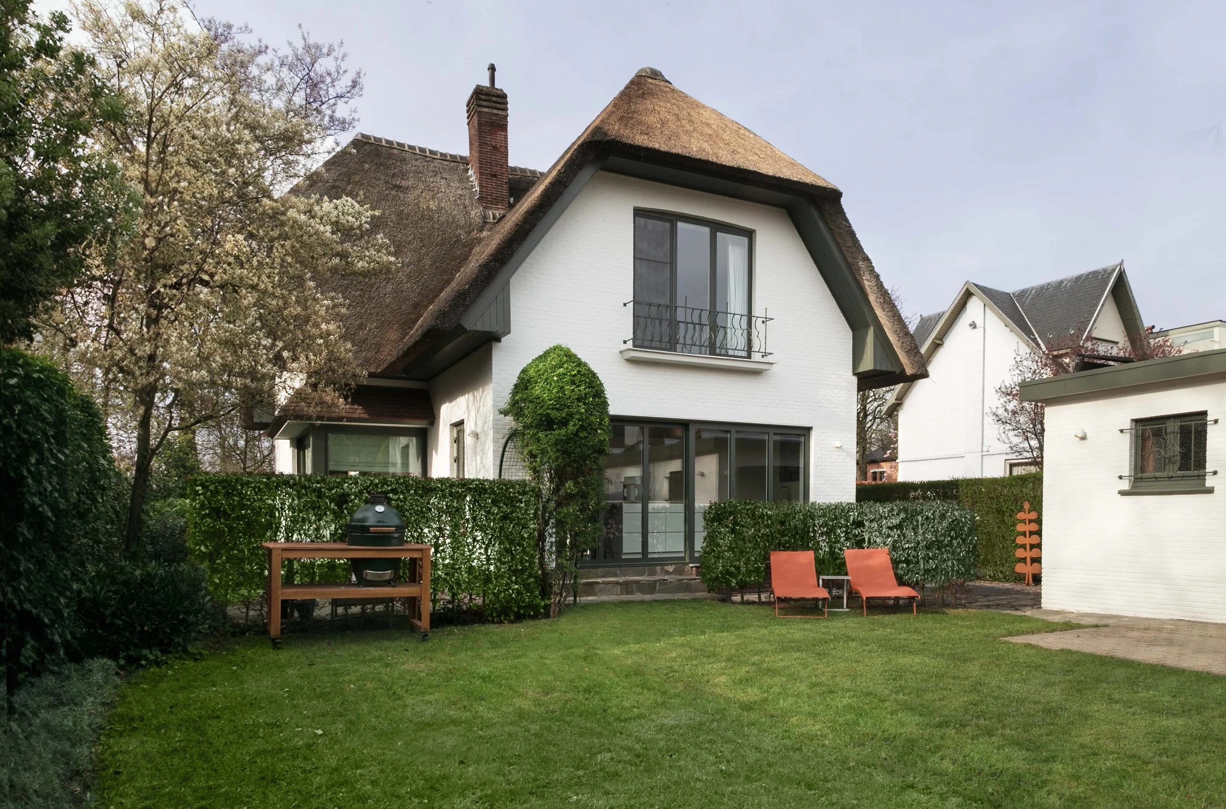 A white modern house with a thatched roof, large glass doors, a small balcony, and surrounding greenery including trees, bushes, and a lawn with outdoor chairs and a barbecue grill.