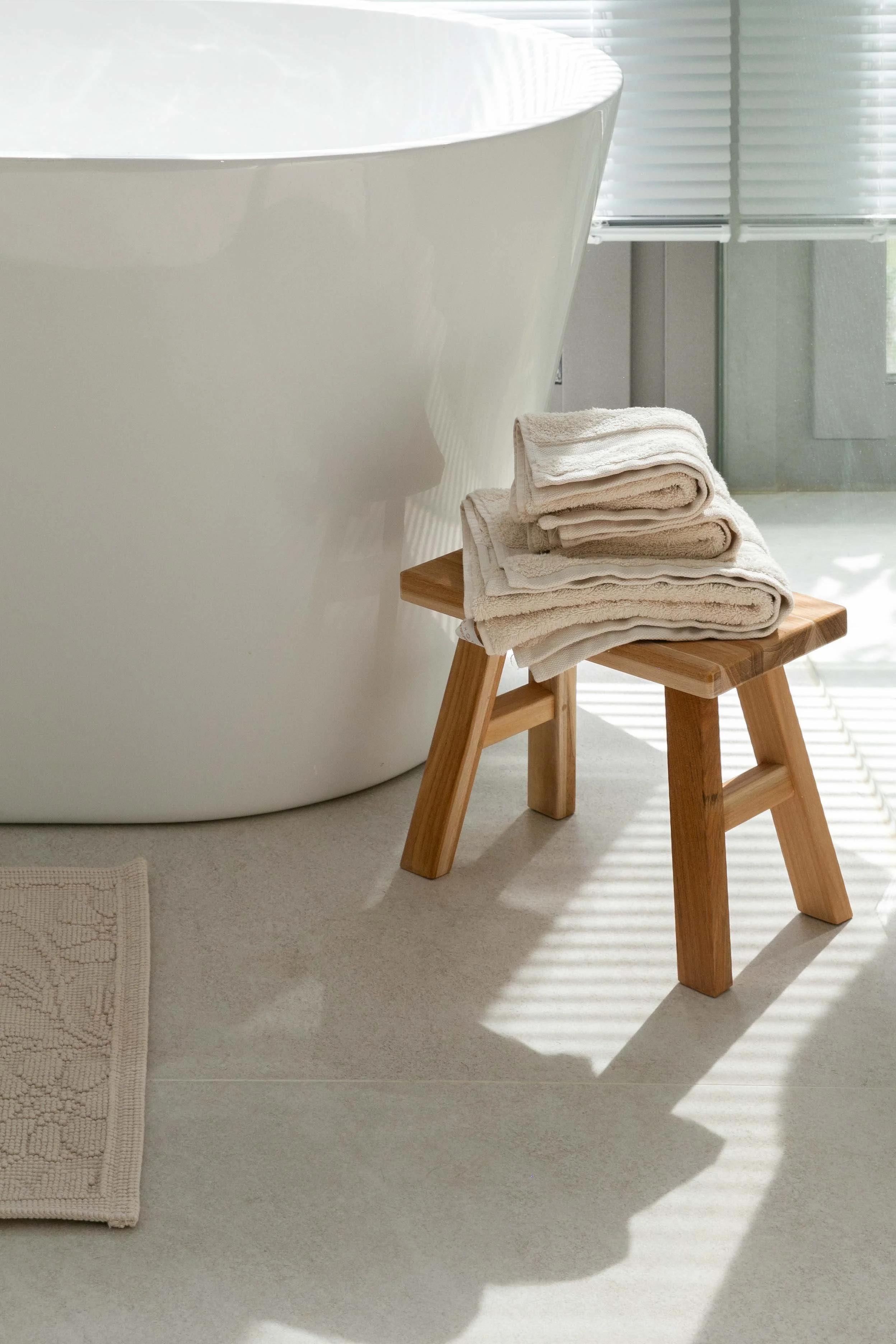 A stack of folded beige and white towels on a small wooden stool next to a white bathtub with a curved side in a modern bathroom with gray flooring and window blinds casting striped shadows.