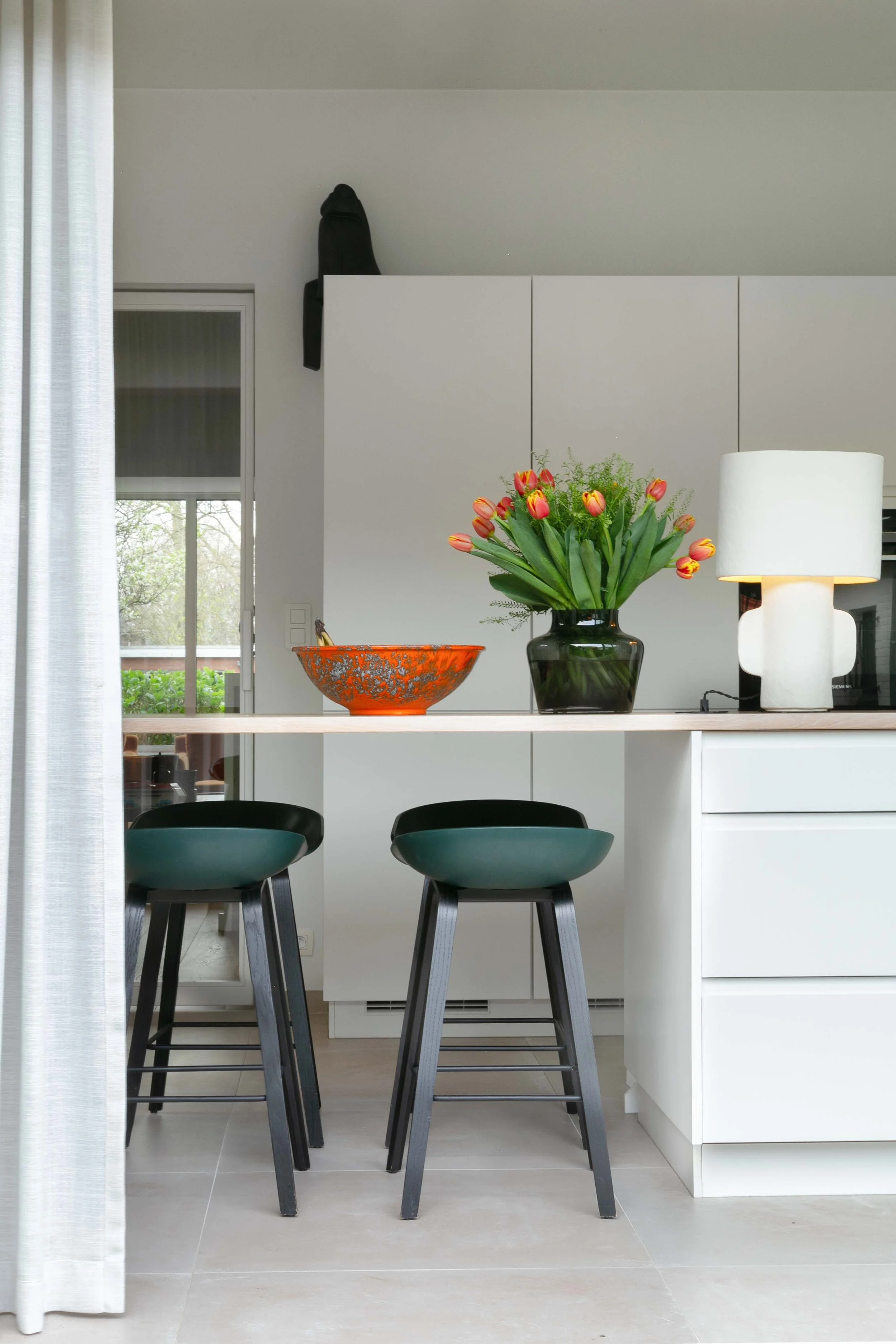 Modern kitchen with white cabinetry, black bar stools, a large white lamp, a vase with pink and yellow tulips, and an orange decorative bowl on a white counter. A sliding glass door and window show greenery outside.