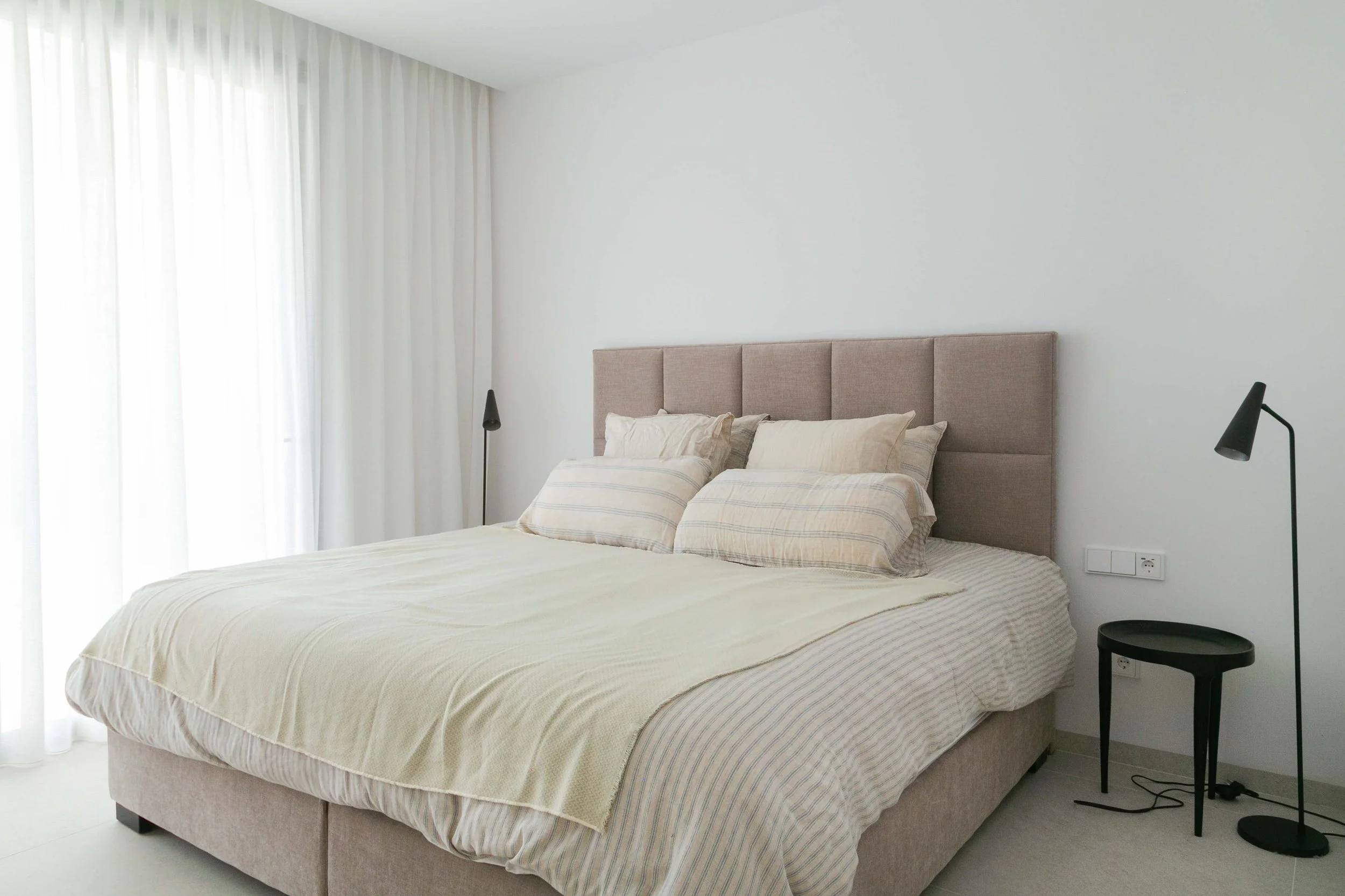 A neatly made bed with striped beige and cream bedding, beige headboard, pillows, bedside lamps, and a black chair with a round side table in a minimalist bedroom with white walls and large window with sheer curtains.