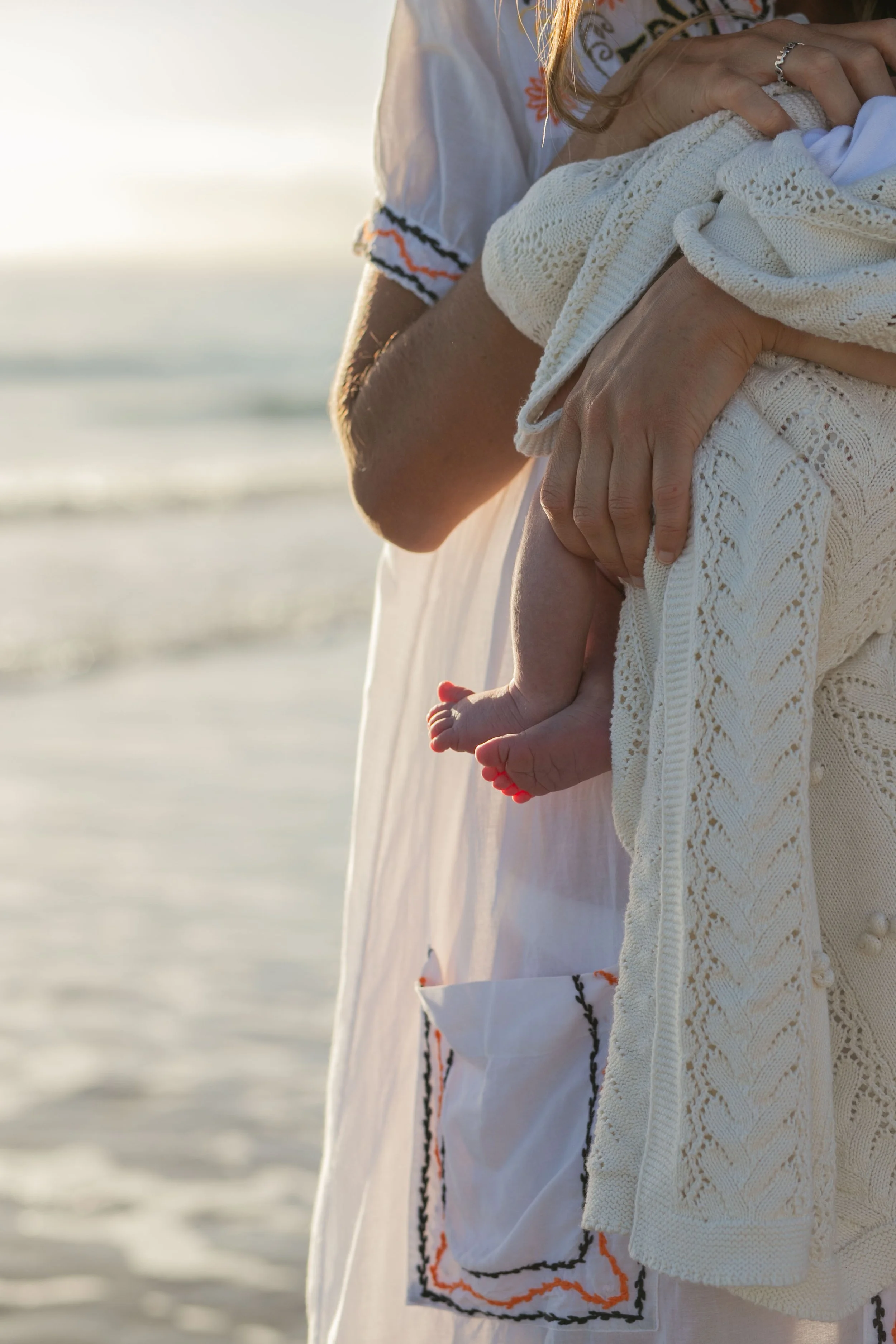 A person holding a small baby wrapped in a cream-colored knit blanket, standing on the beach during sunset.