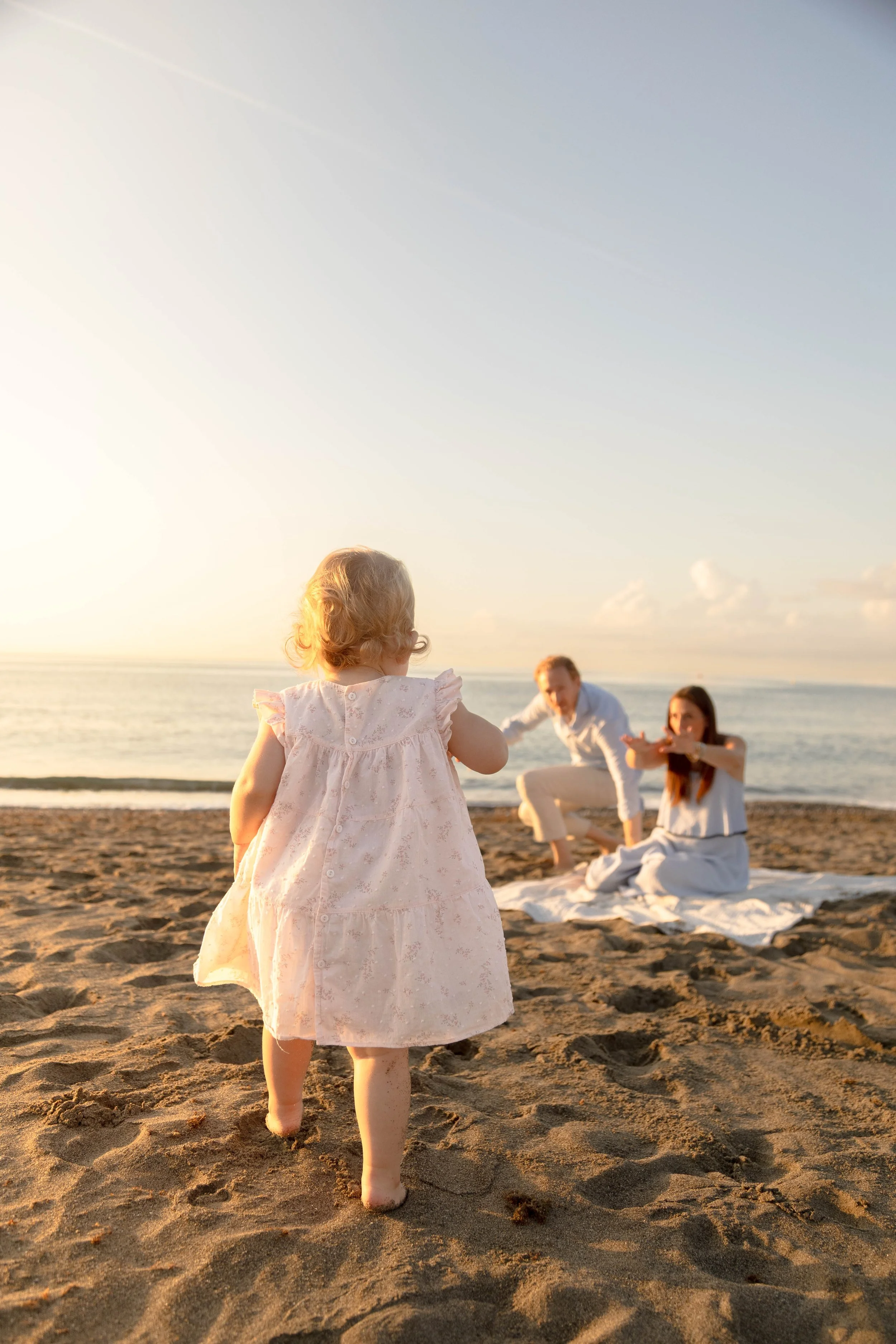 A young girl with blonde hair wearing a light pink dress walking barefoot on the sandy beach towards a family sitting on a white blanket, with the ocean and a sunset sky in the background.
