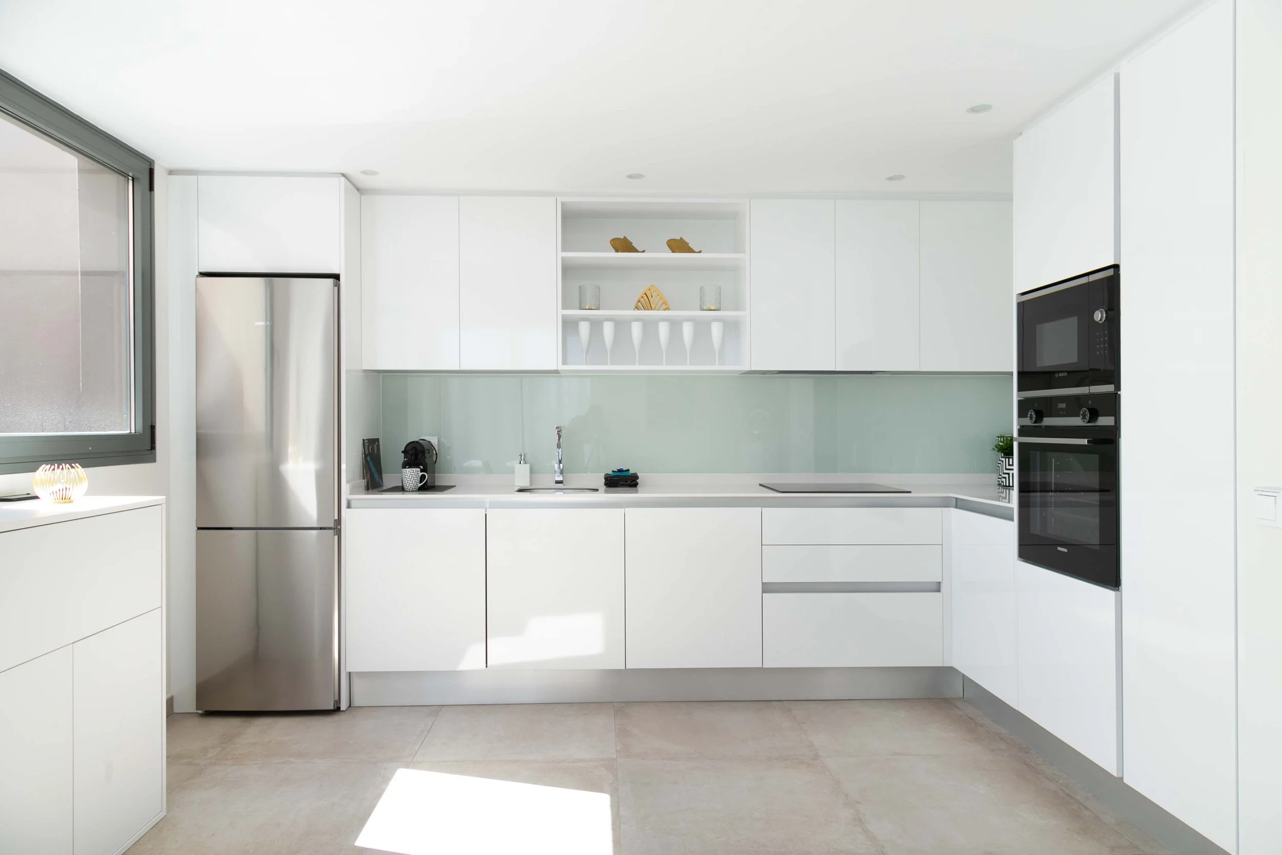 Modern white kitchen with built-in appliances, open shelving with decorative items, and a window letting in natural light.
