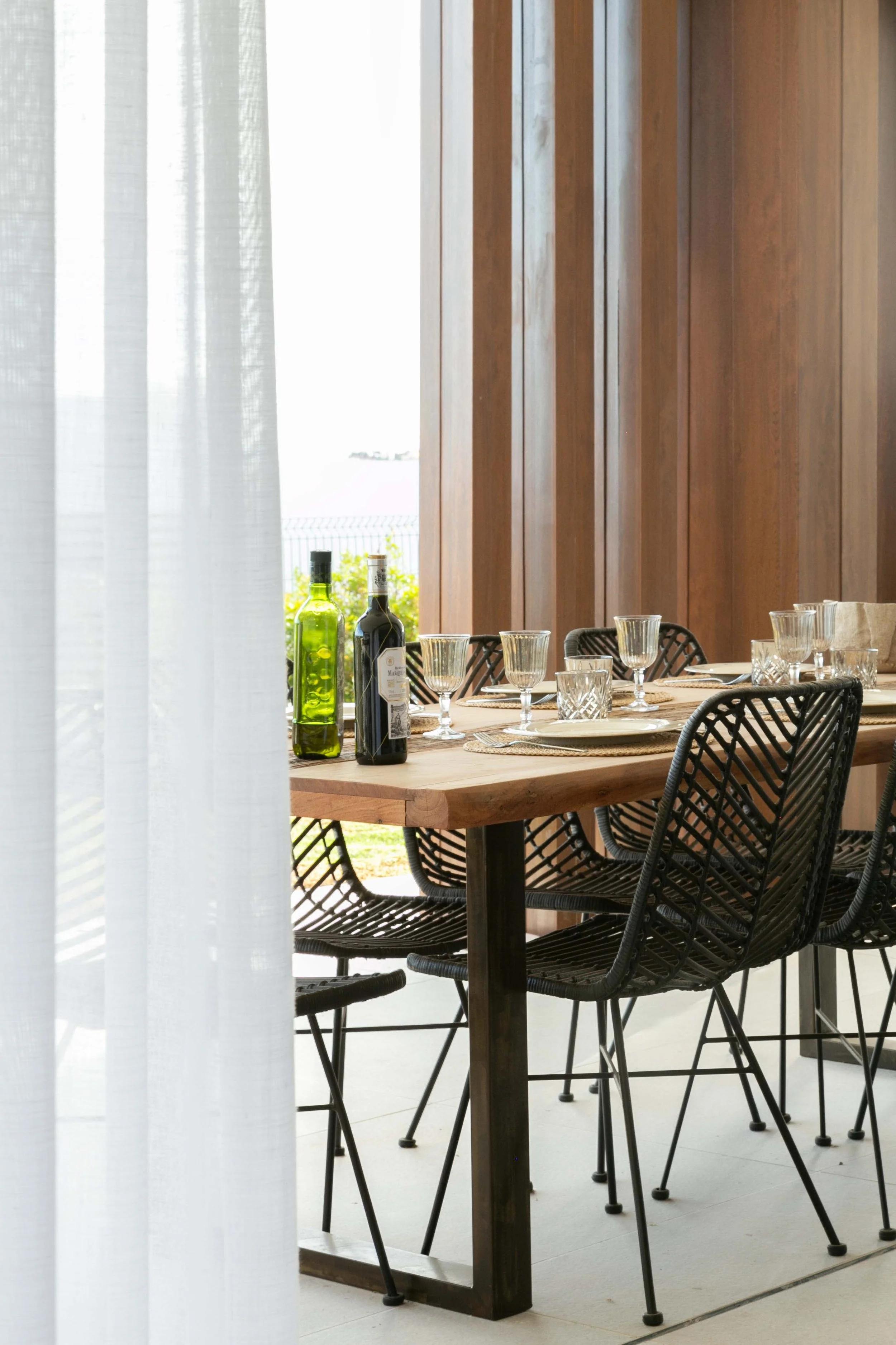 A dining table set with glasses, plates, and bottles in a room with wooden paneling and sheer curtains.