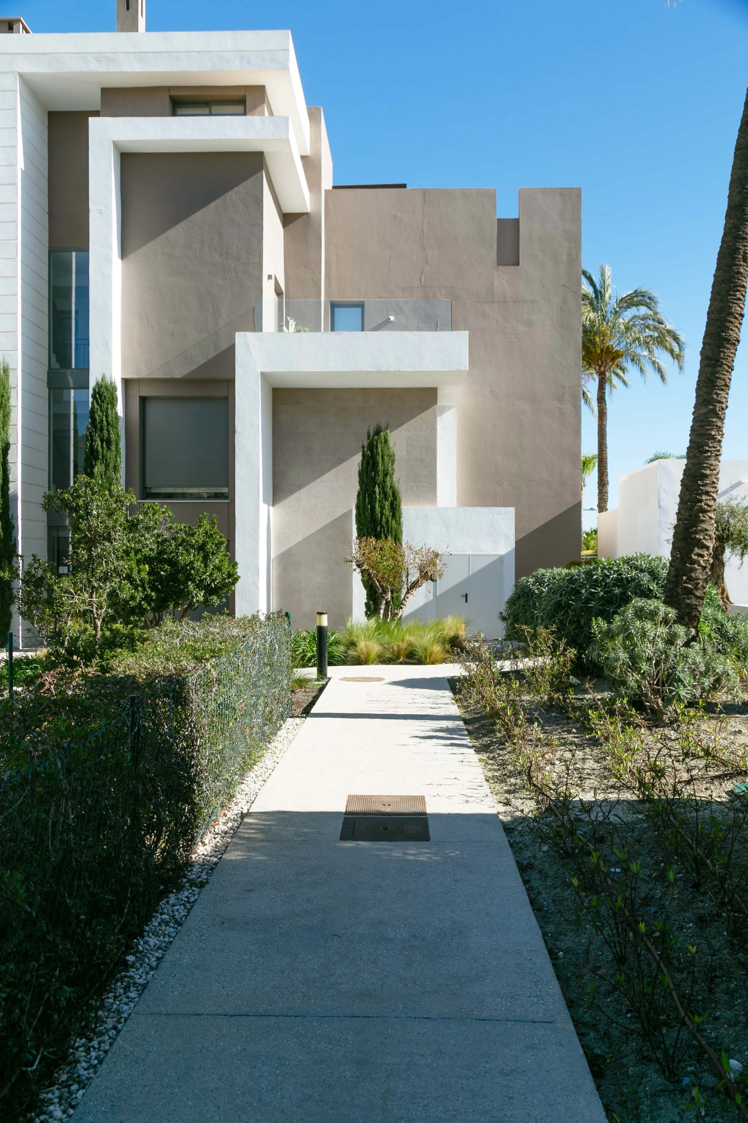 Modern residential building with white and beige walls, glass balcony, and a pathway surrounded by green bushes and palm trees under a bright blue sky.