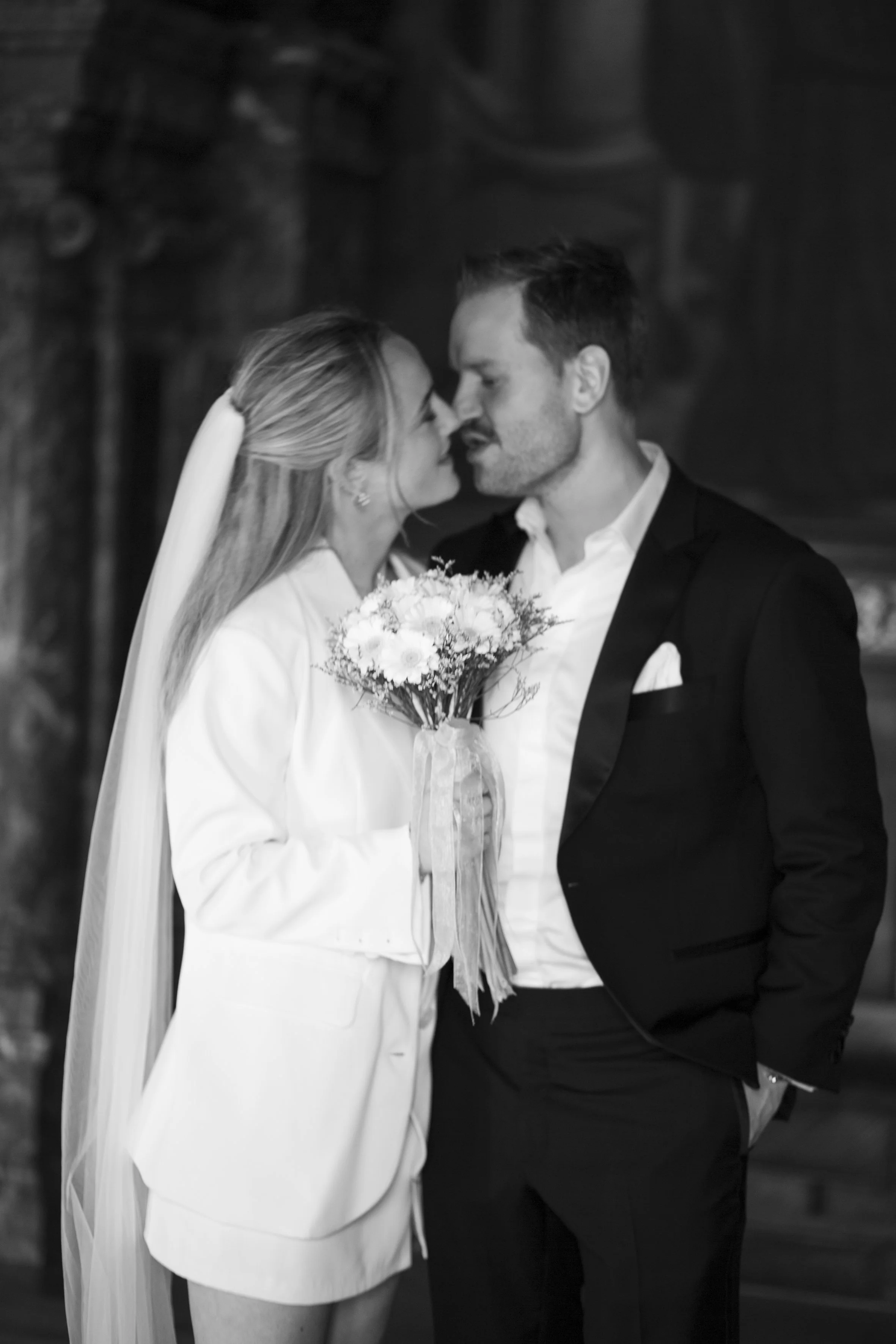 Black and white photo of a bride and groom standing close, with their foreheads touching, holding a bouquet of flowers. The bride wears a white dress with a veil, and the groom wears a suit with an open-collared shirt.