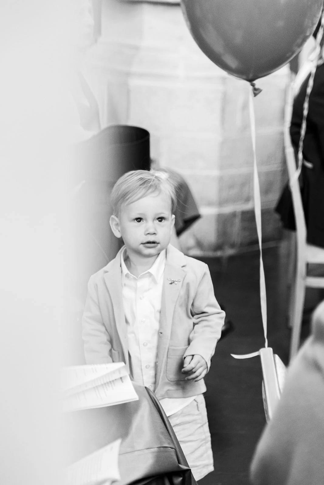 A young boy with light-colored hair wearing a light-colored blazer and shirt, standing next to a table with open books or papers, in a room decorated for a celebration with balloons, including a large dark balloon, nearby. The scene appears to be ind