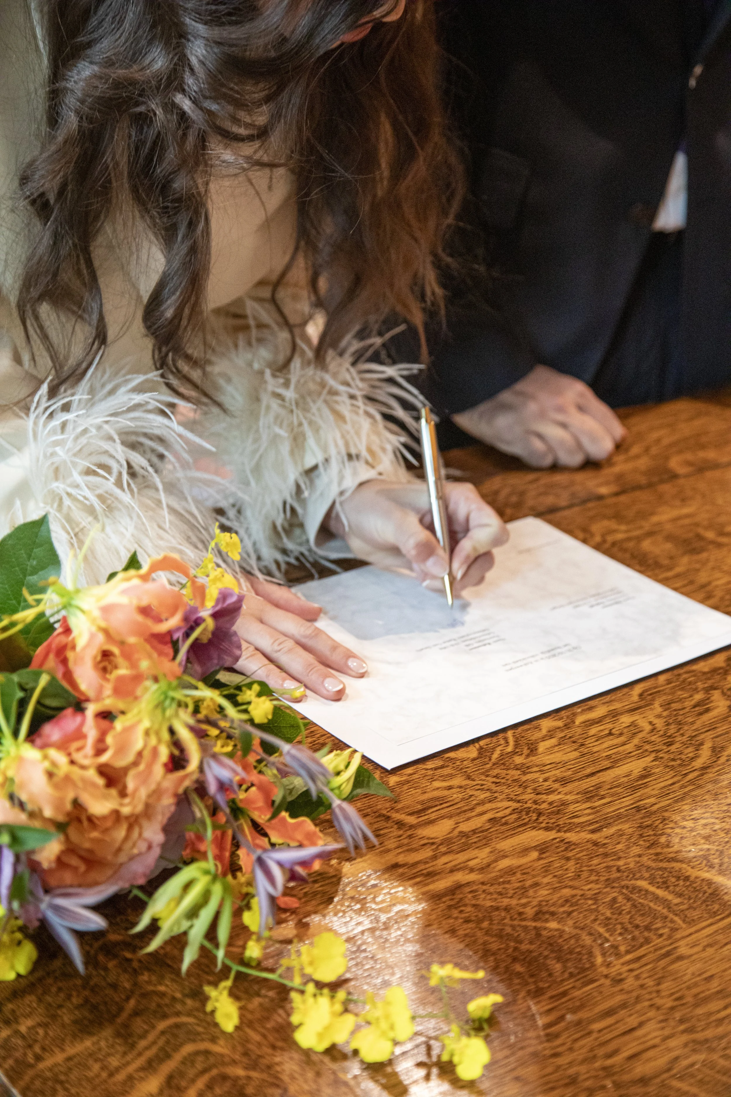 A woman with curly brown hair signs a document on a wooden table next to a colorful bouquet of flowers.