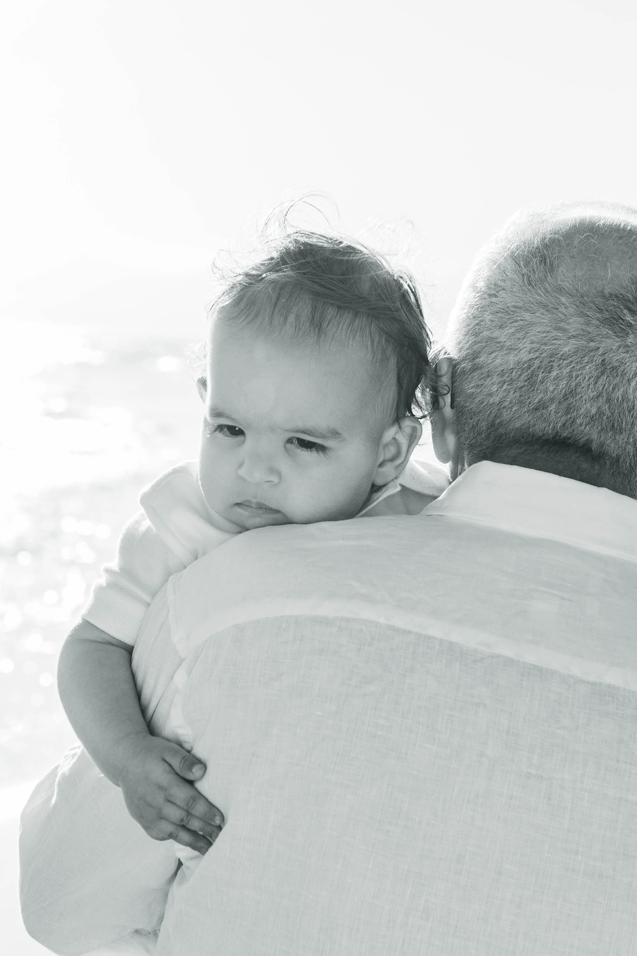 A young child with puffy hair being held by an adult. The child looks slightly to the side with a neutral expression. The adult's back is visible, showing a short gray haircut and a light-colored shirt. The background appears to be an outdoor setting