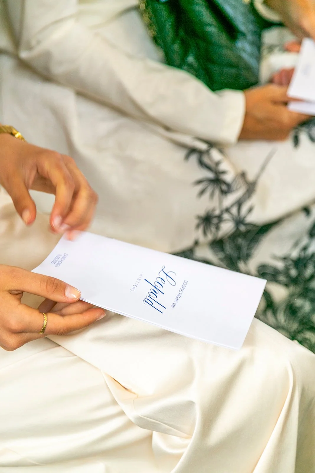 A person holding a white wedding invitation with elegant blue script that reads 'We Do! Love' as another individual in the background holds a magazine or program and a green plant.