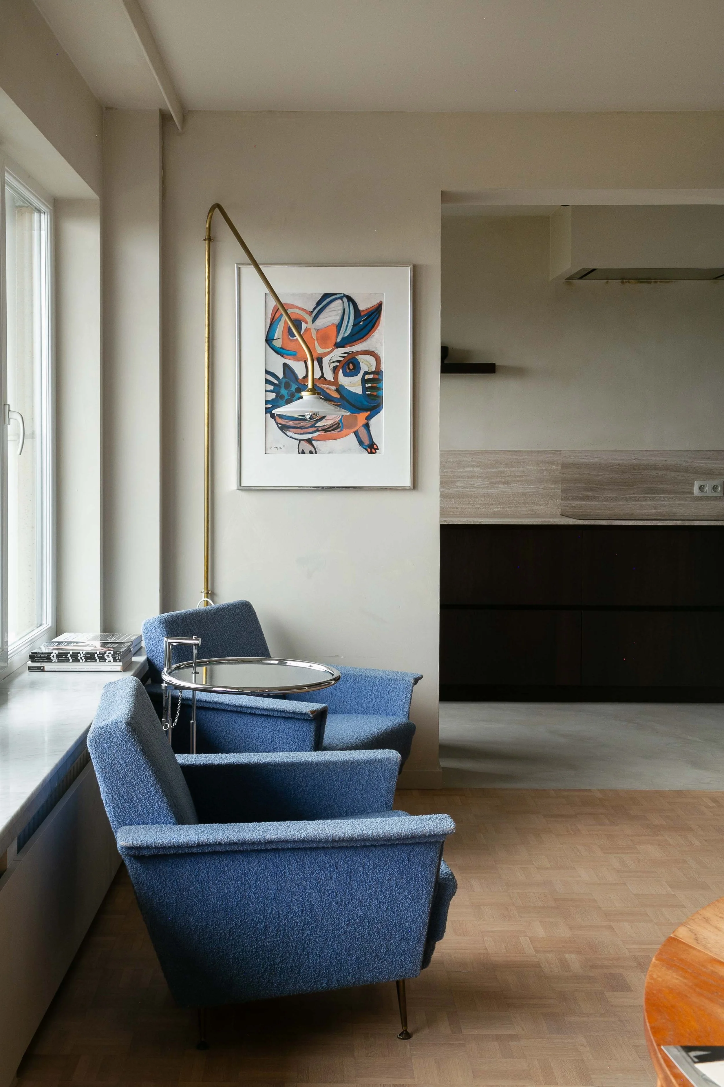 Interior of a room with blue upholstered chairs, a table with magazines, a framed abstract art piece on the wall, and a large window letting in natural light.