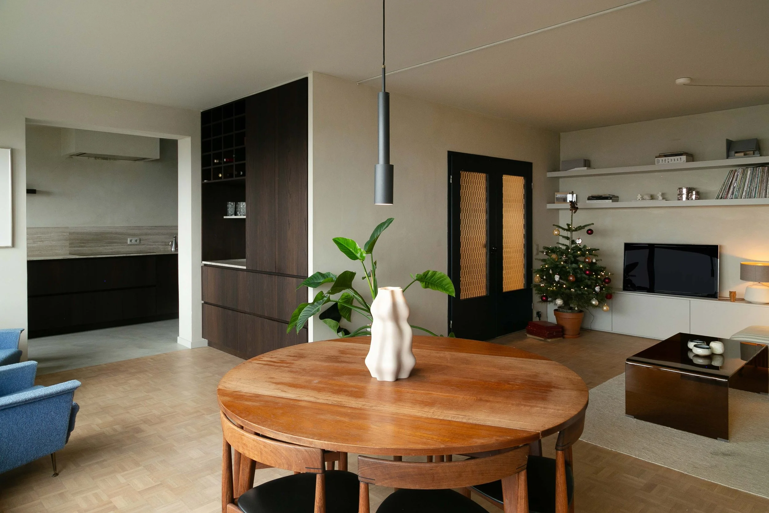 Living room with a round wooden dining table, a decorative white vase with green leaves, a small decorated Christmas tree, a blue armchair, a black door with glass panels, and a wall-mounted white shelf holding books and decorative items.
