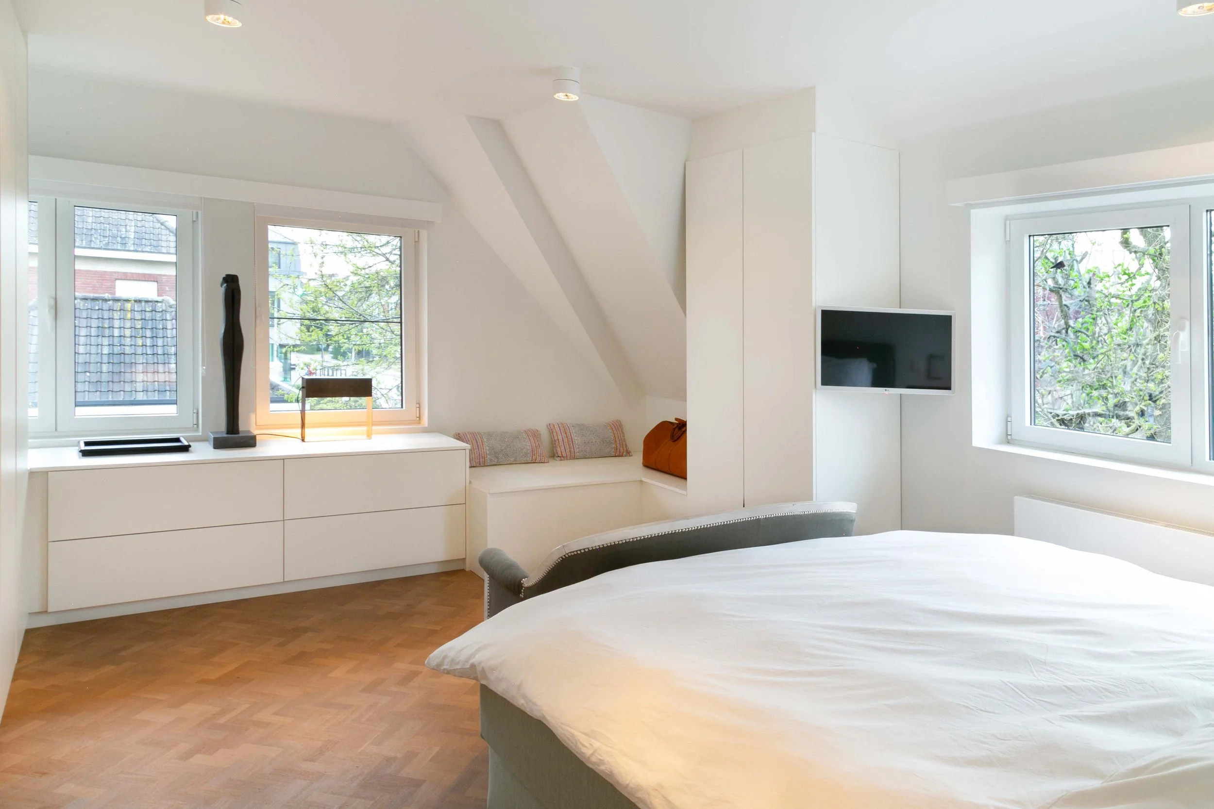 A bedroom with white walls and a sloped ceiling, featuring two windows with trees outside. There is a bed with white bedding in the foreground, and a built-in window seat with cushions and decorative items on the window sill. A flat-screen TV is moun