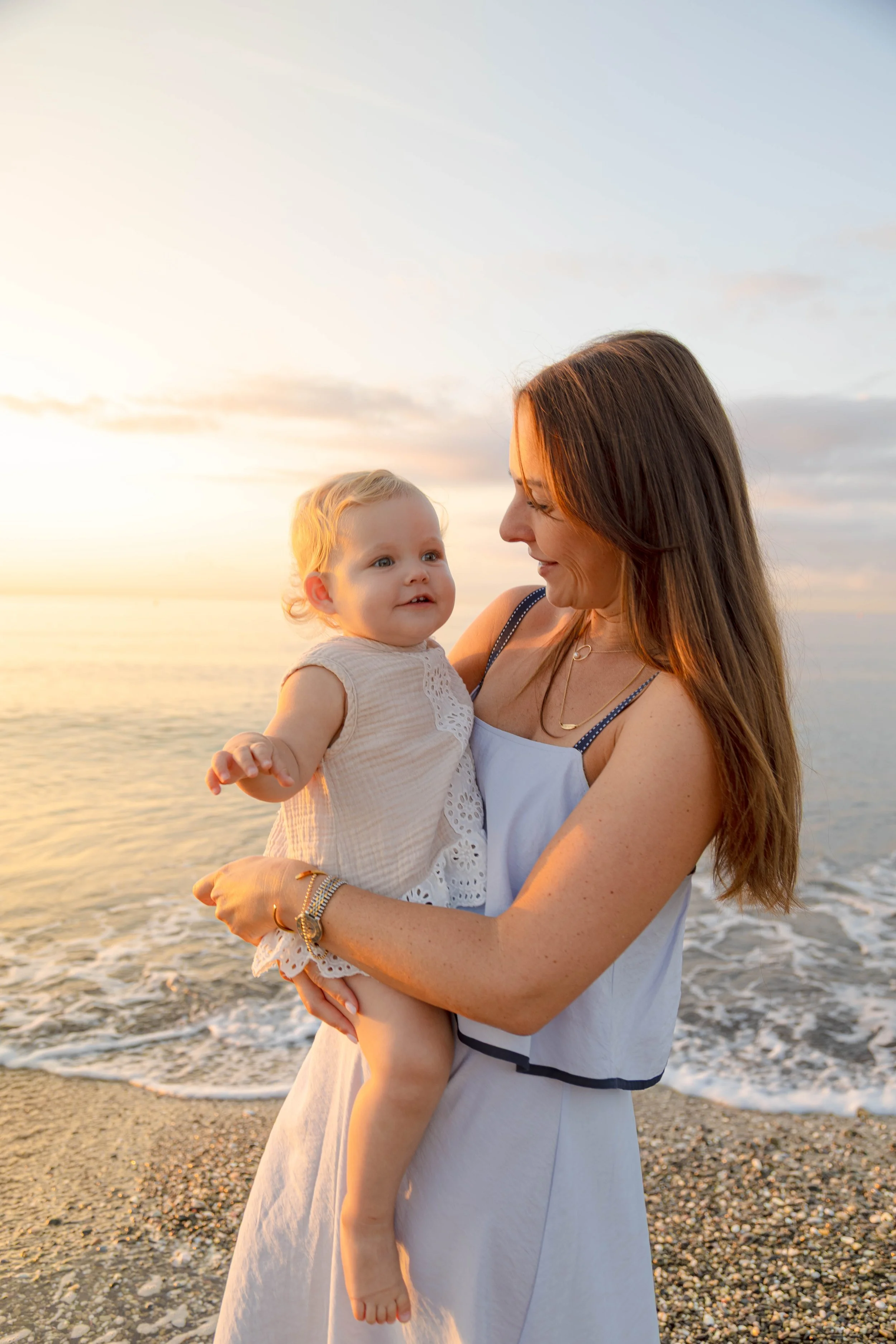 A mother holding a baby at the beach during sunset, with the ocean and cloudy sky in the background. Golden hour photography marbella