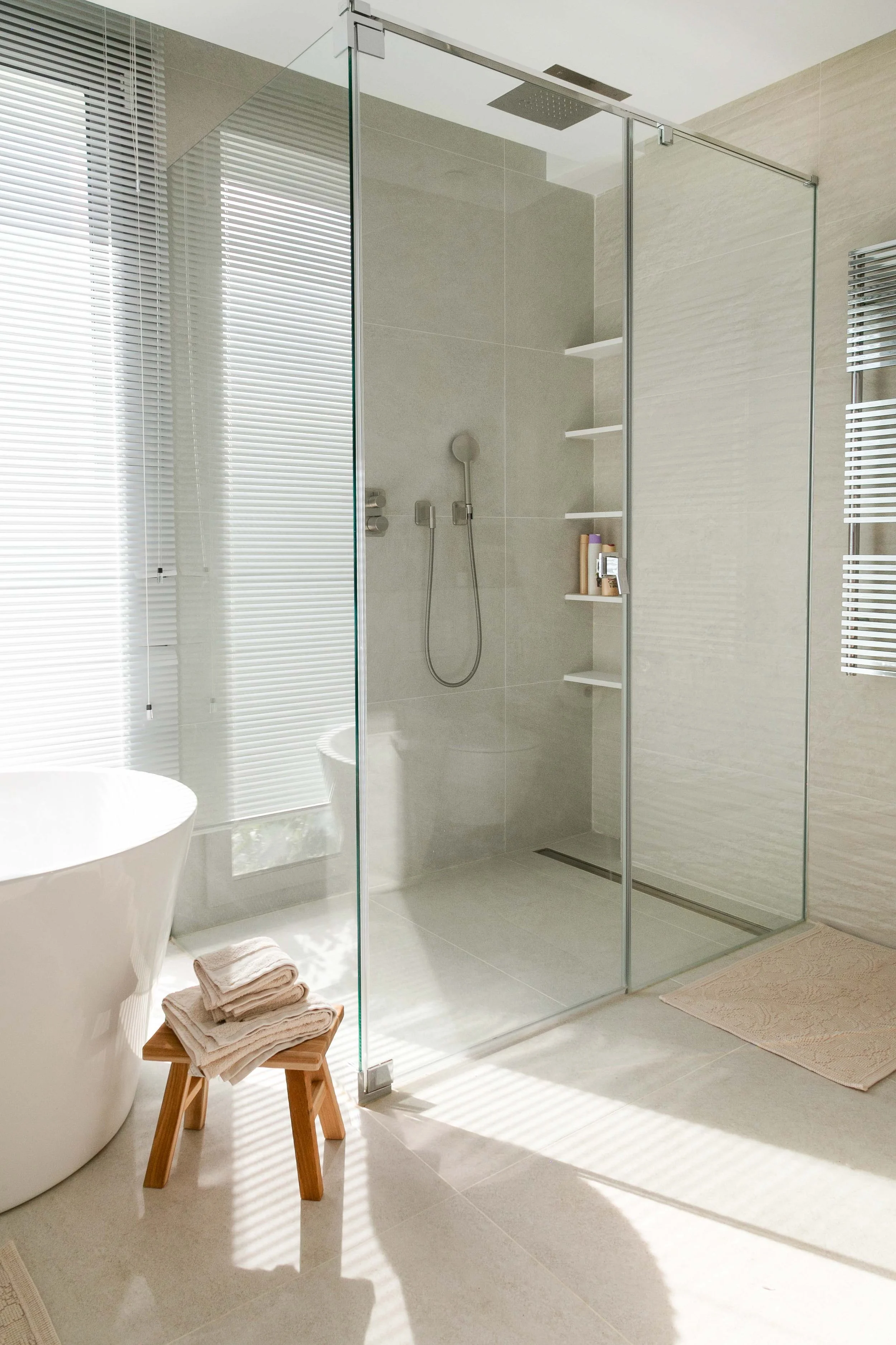 A modern bathroom with a glass-enclosed shower, a white freestanding bathtub, a small wooden stool with folded towels, and natural light coming through blinds.