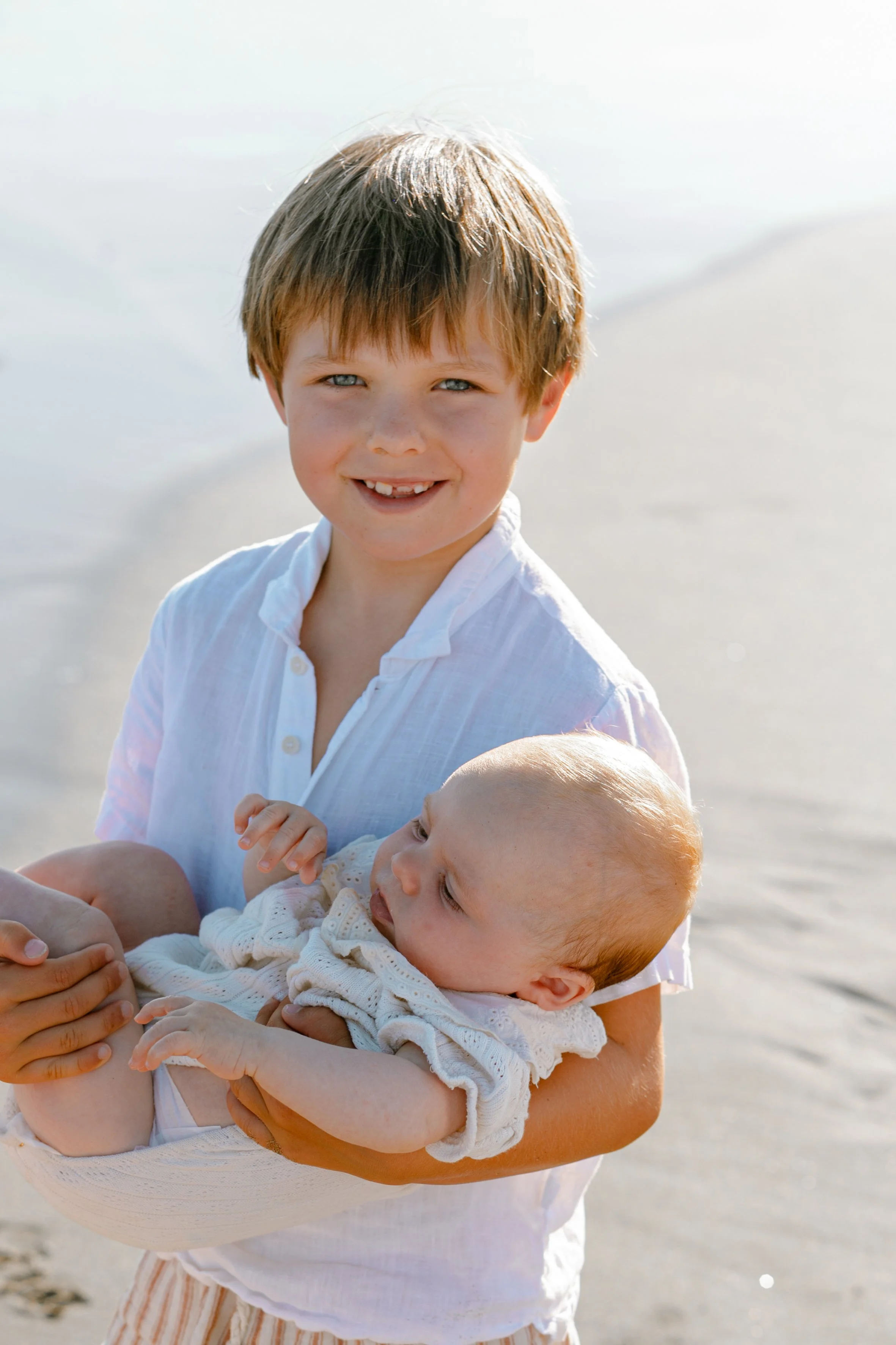 A young boy with brown hair and blue eyes holding a baby with light hair on a beach.