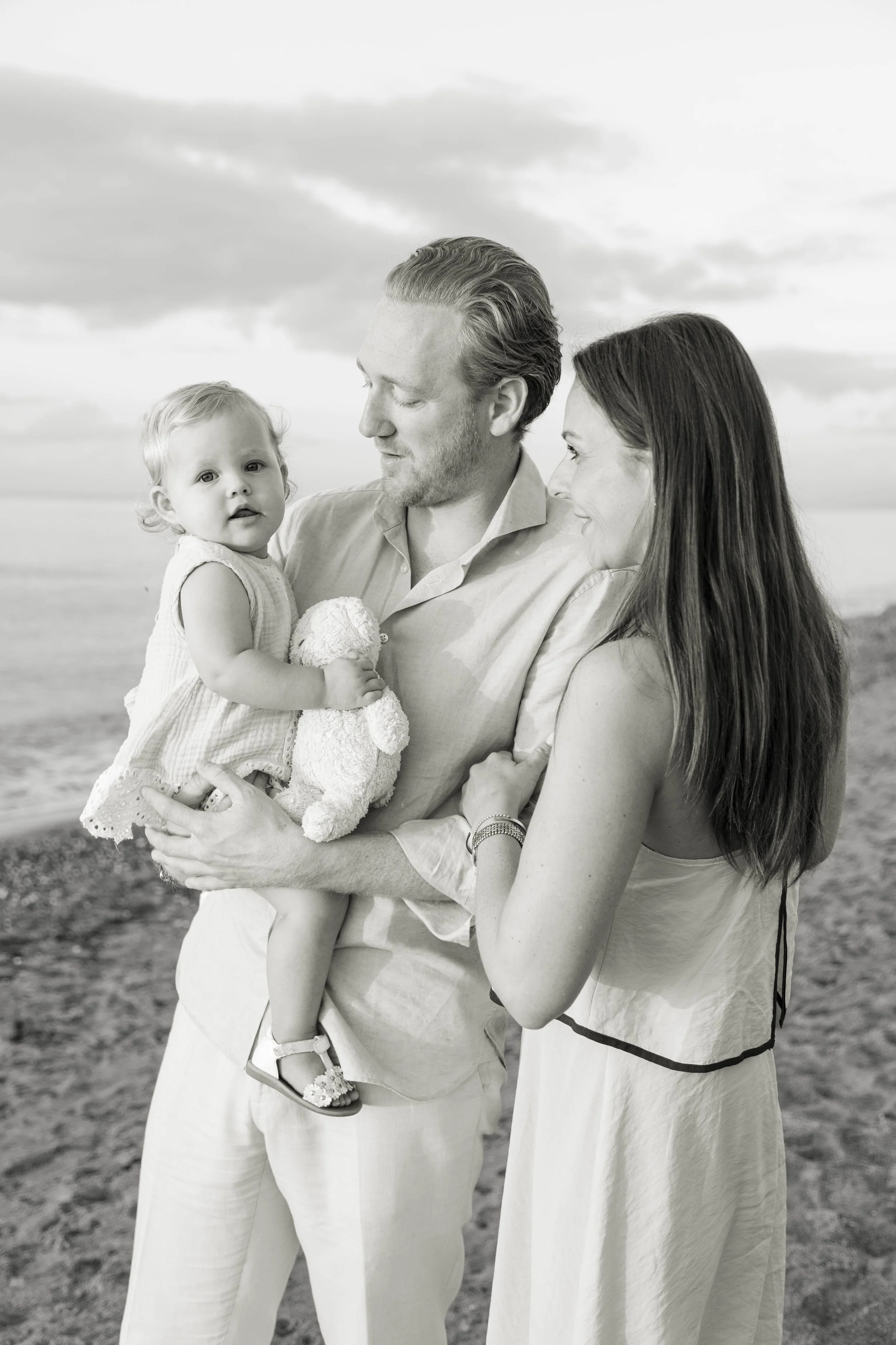 A black and white photo of a family outdoors at the beach, with a man holding a young girl and a woman standing close beside him, all smiling and looking at each other.