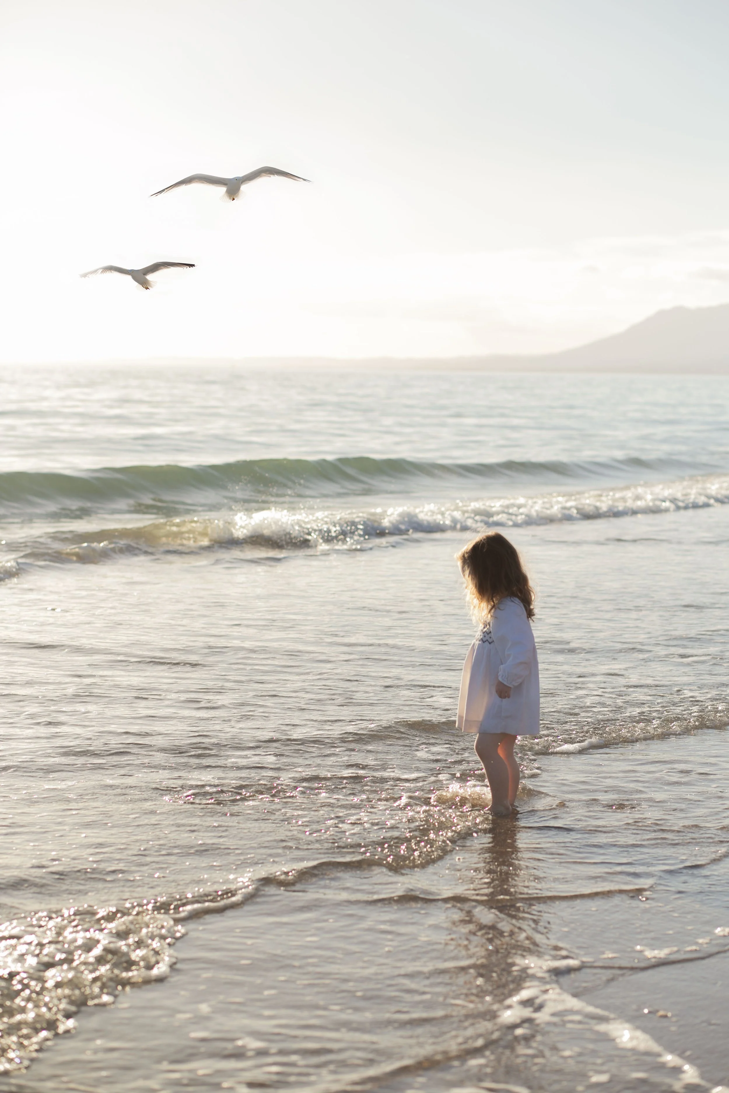Child standing in the shallow ocean water near the shore, looking at seagulls flying above, with mountains in the background, during sunset or sunrise. Photography for a child in marbella on golden hour
