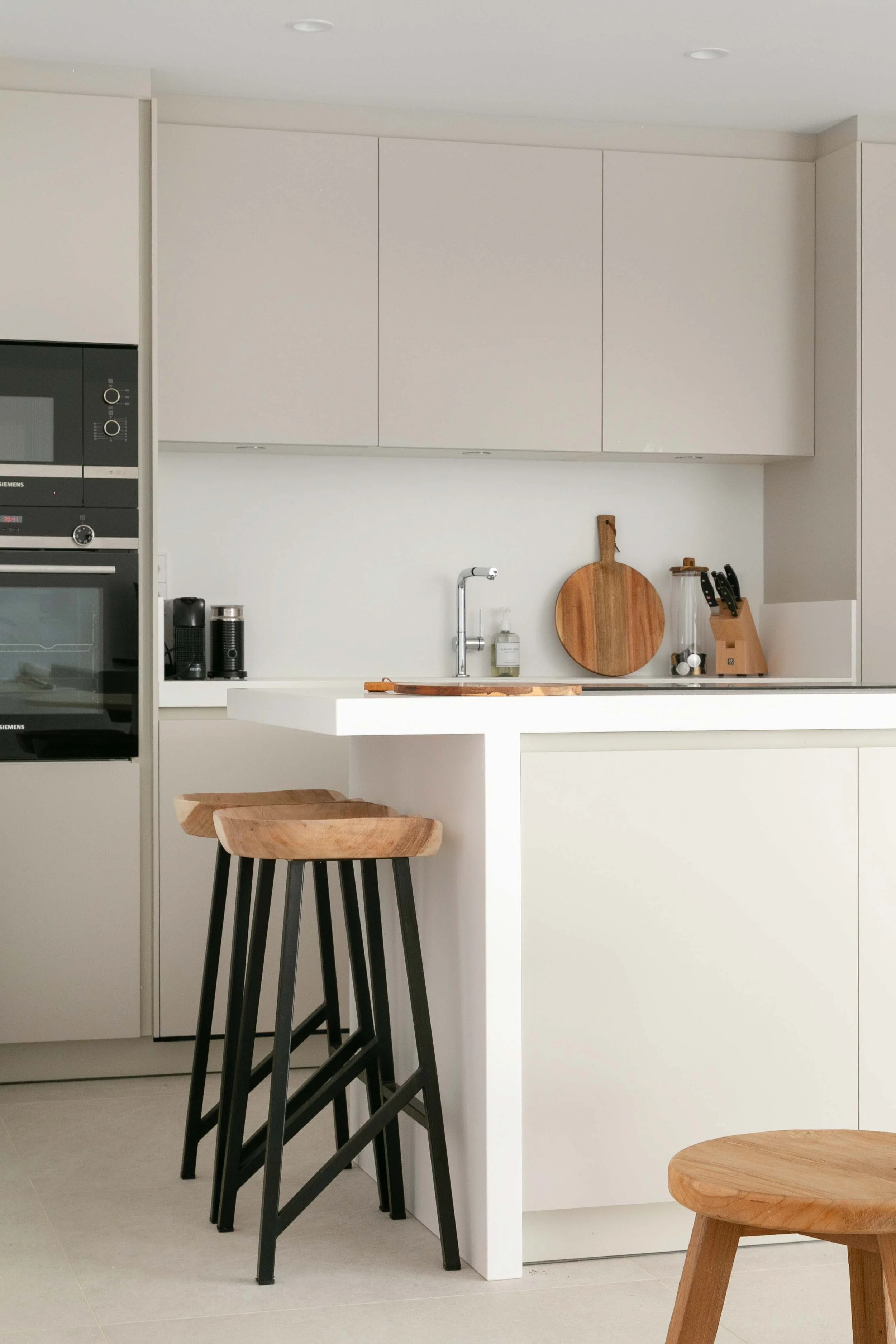 Modern kitchen with white cabinetry, wooden bar stools, black chairs, a round wooden cutting board, and kitchen appliances.