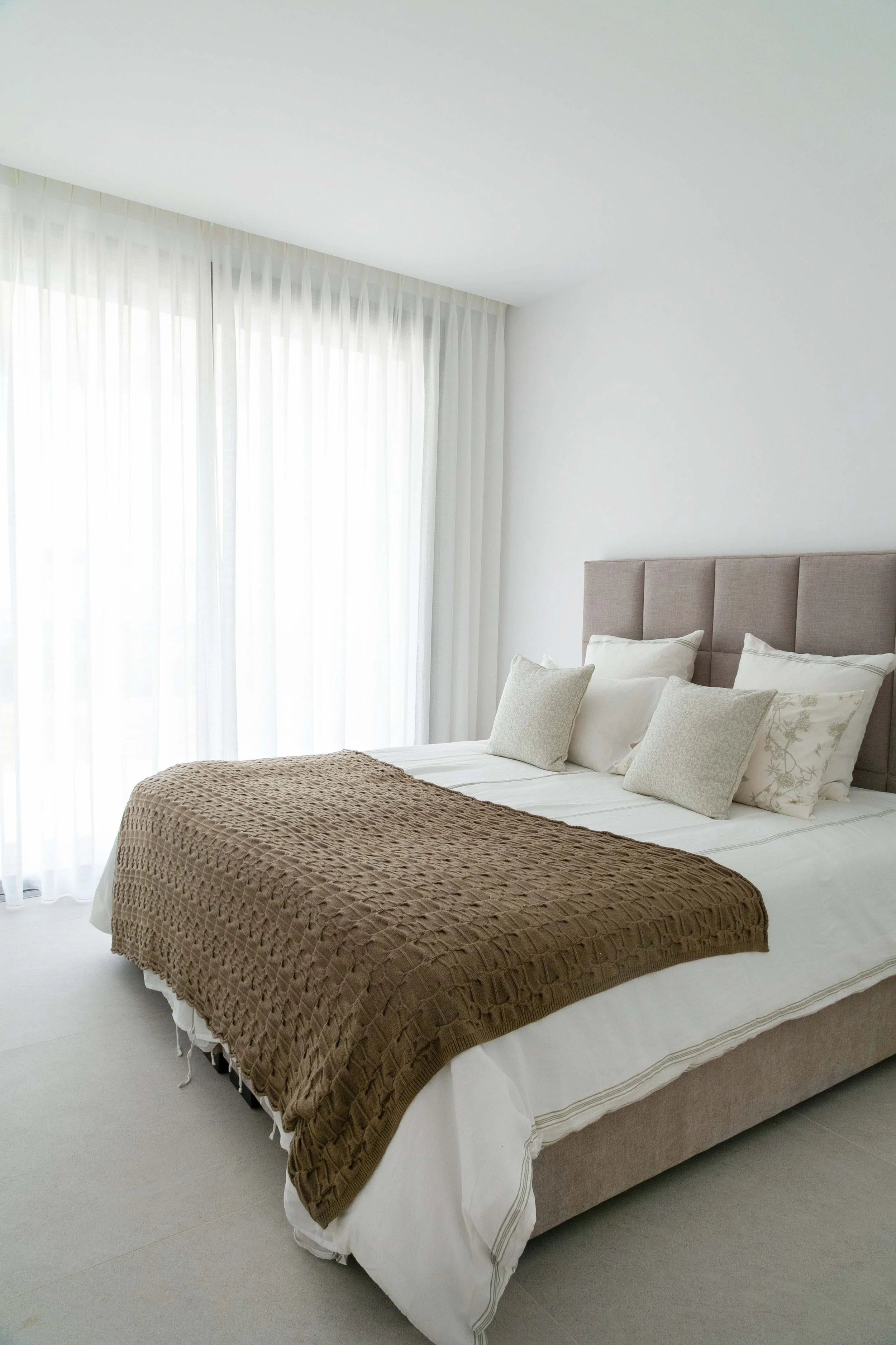 A neatly made bed with white sheets, multiple pillows, and a brown textured blanket in a bright, minimalist bedroom with white walls and large windows covered by sheer white curtains.