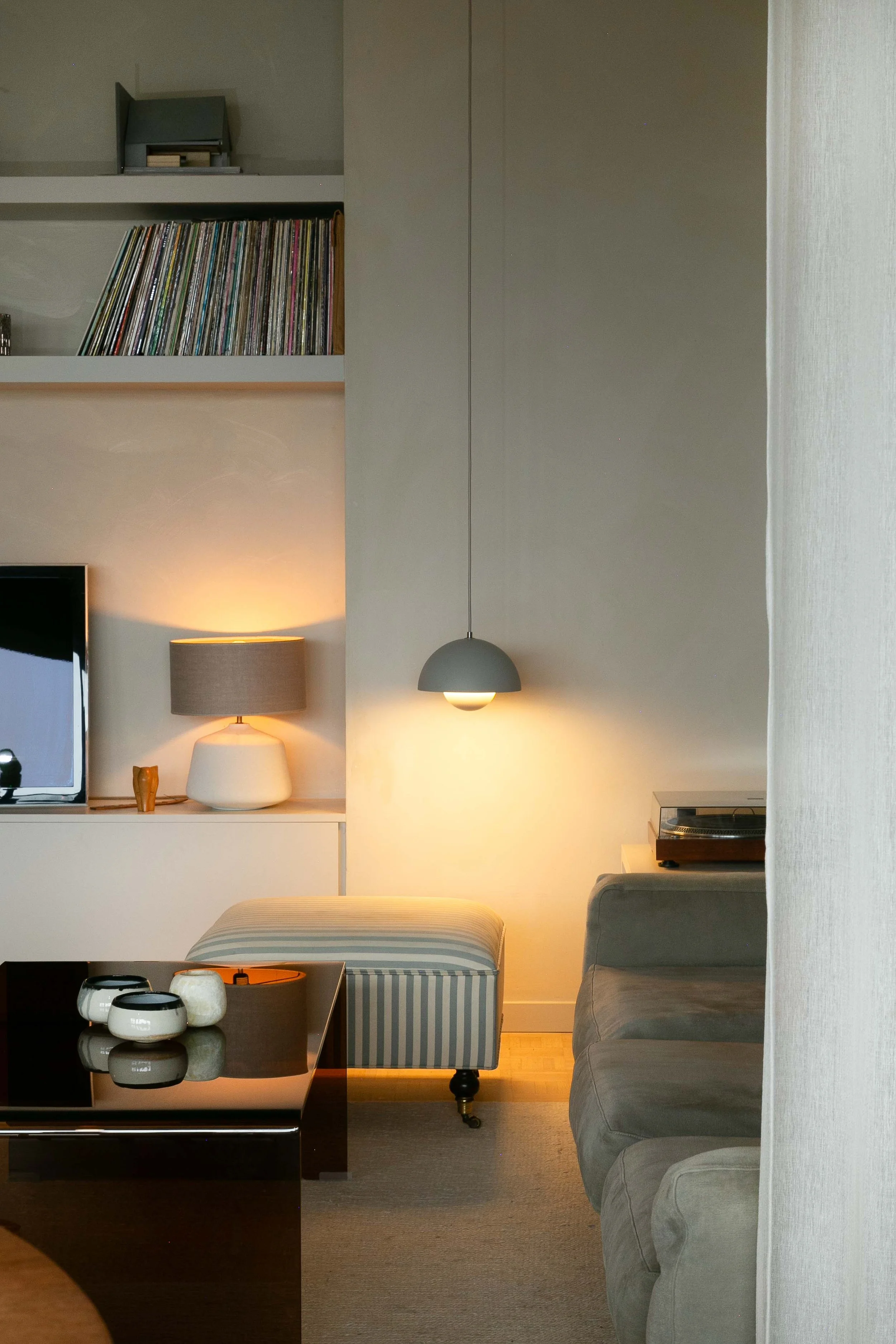 A cozy living room with a television, a table with decorative bowls, a striped upholstered stool, a gray sofa, and a wall with a shelf holding vinyl records. There are two lamps providing warm lighting, and part of a white curtain on the right.