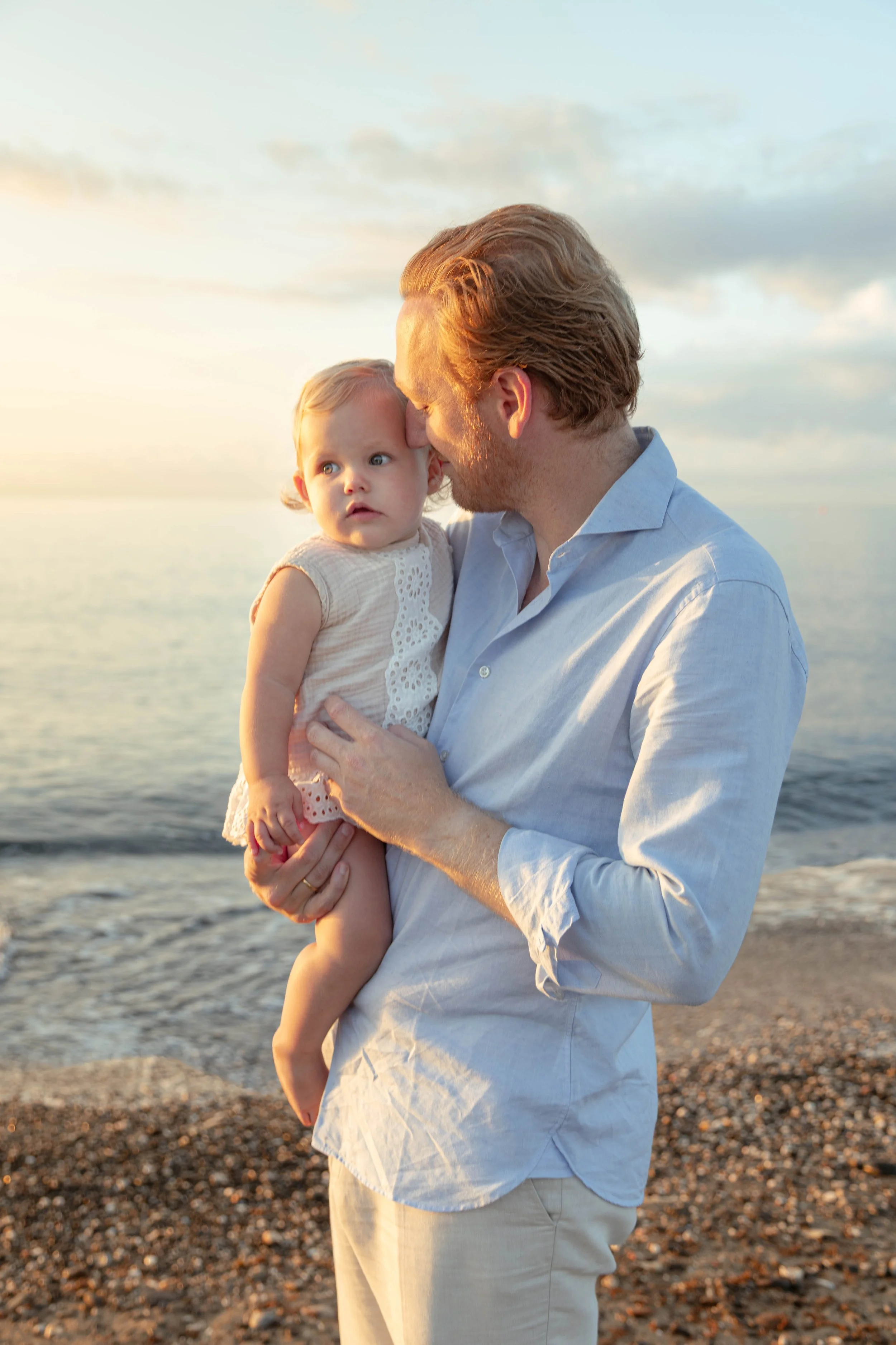 A man holding a young girl at the beach during sunset, with the ocean and cloudy sky in the background.