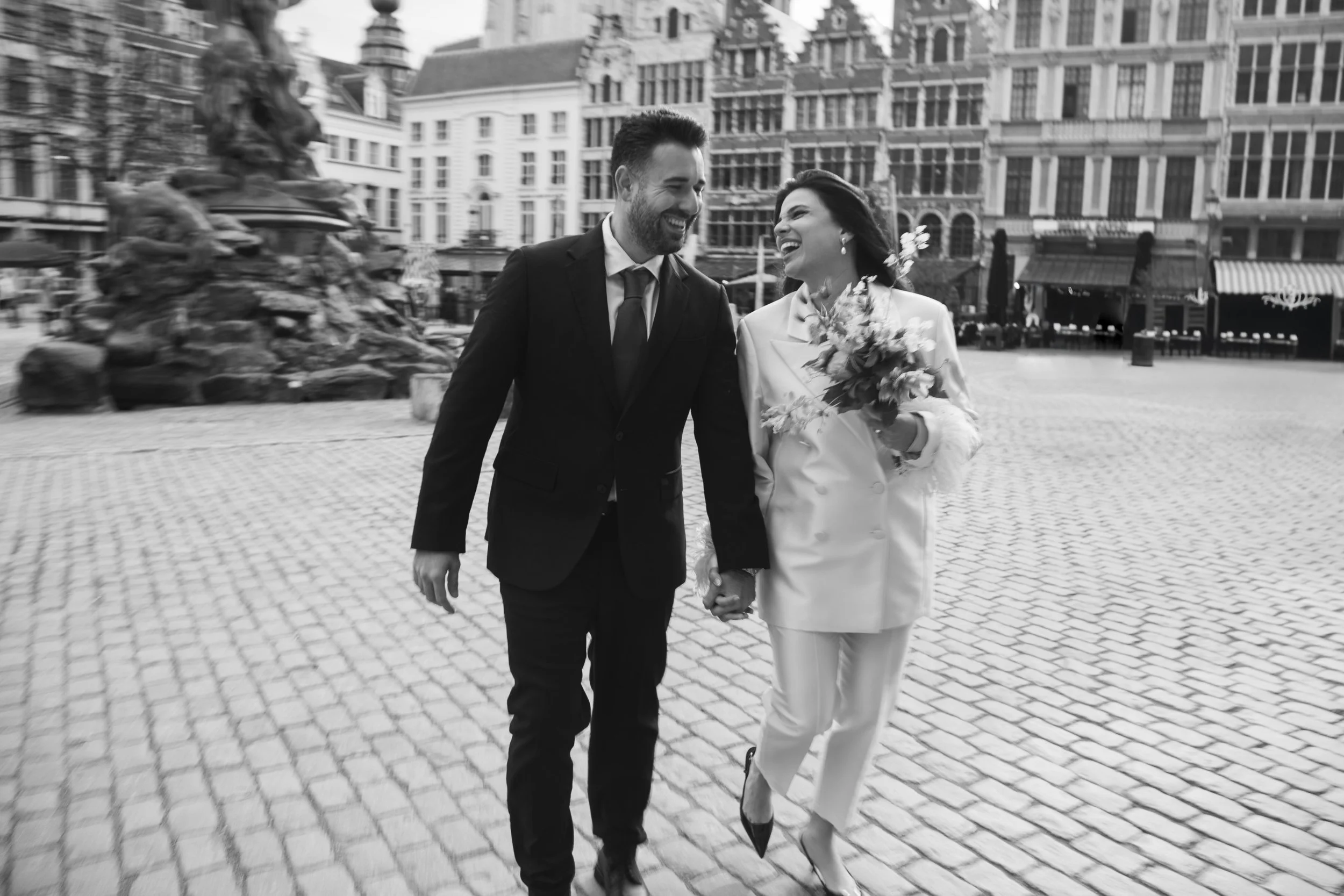 A happy couple holding hands and smiling as they walk together in an urban plaza. The woman holds a bouquet of flowers and wears a white blazer with matching pants, while the man is dressed in a black suit and tie.