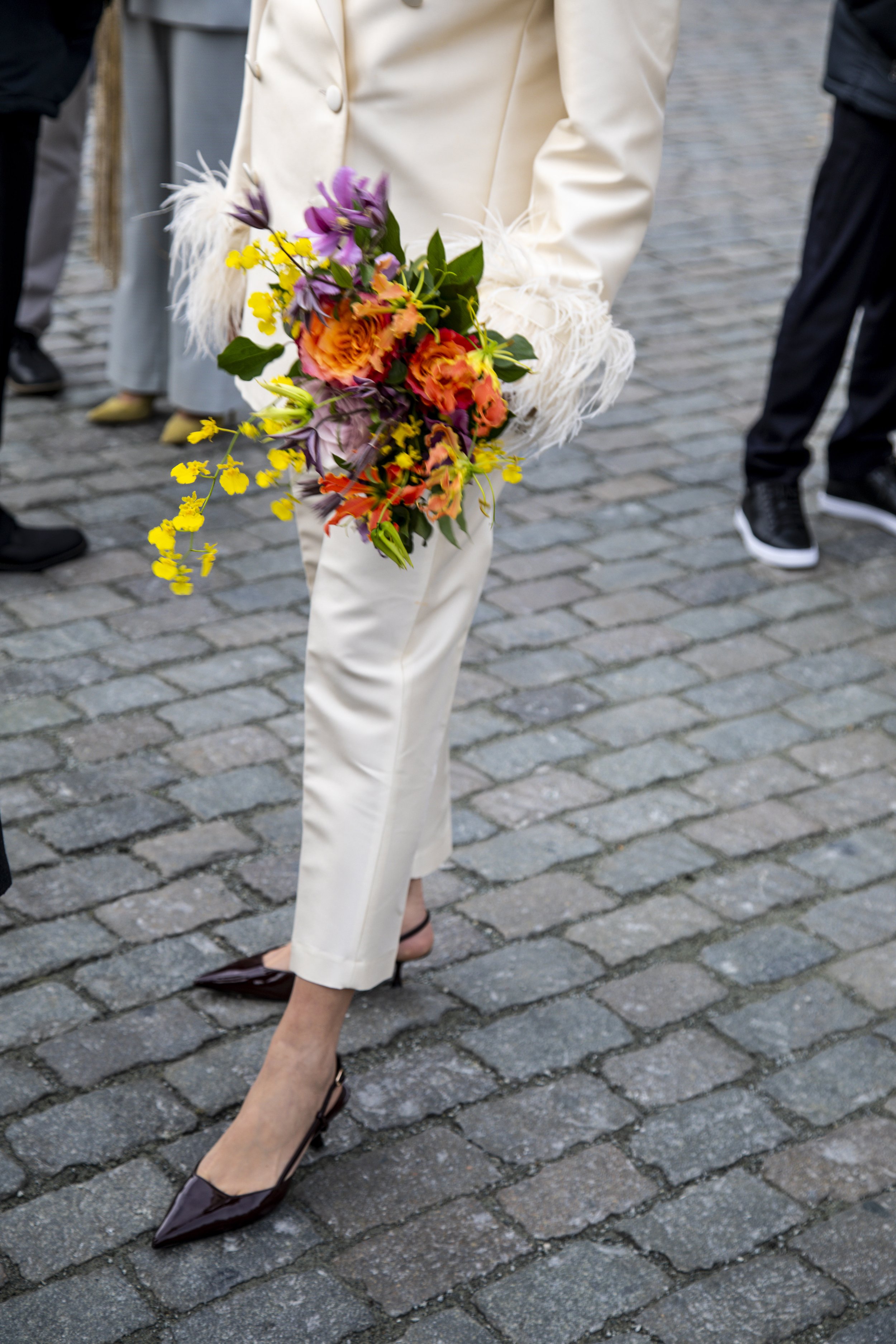 Person standing on cobblestone street holding a colorful bouquet of flowers, wearing cream-colored pants, high heels, and a white jacket with feathered cuffs, surrounded by other people in casual attire.